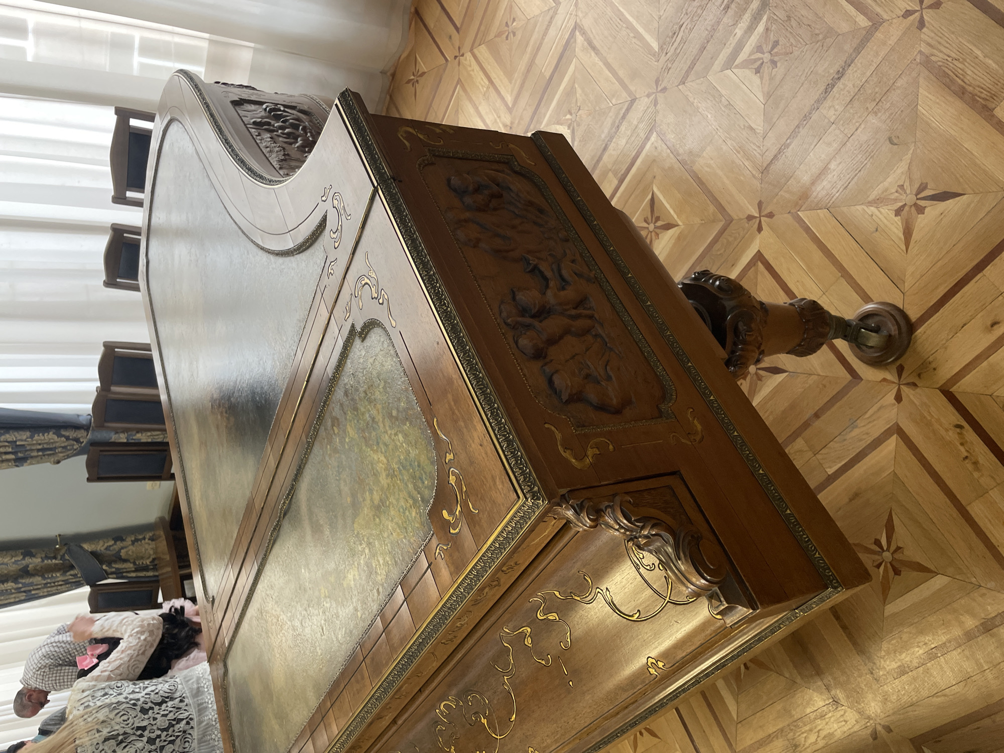 The image shows a detailed view of an ornate, antique wooden cabinet in a room with parquet flooring. The cabinet is decorated with carvings, molding, and gold accents. In the background, partly visible through what appears to be a reflective or mirrored surface of the cabinet, are people and furniture, suggesting an interior setting. Based on visual cues, this is located in Yerevan, Armenia.