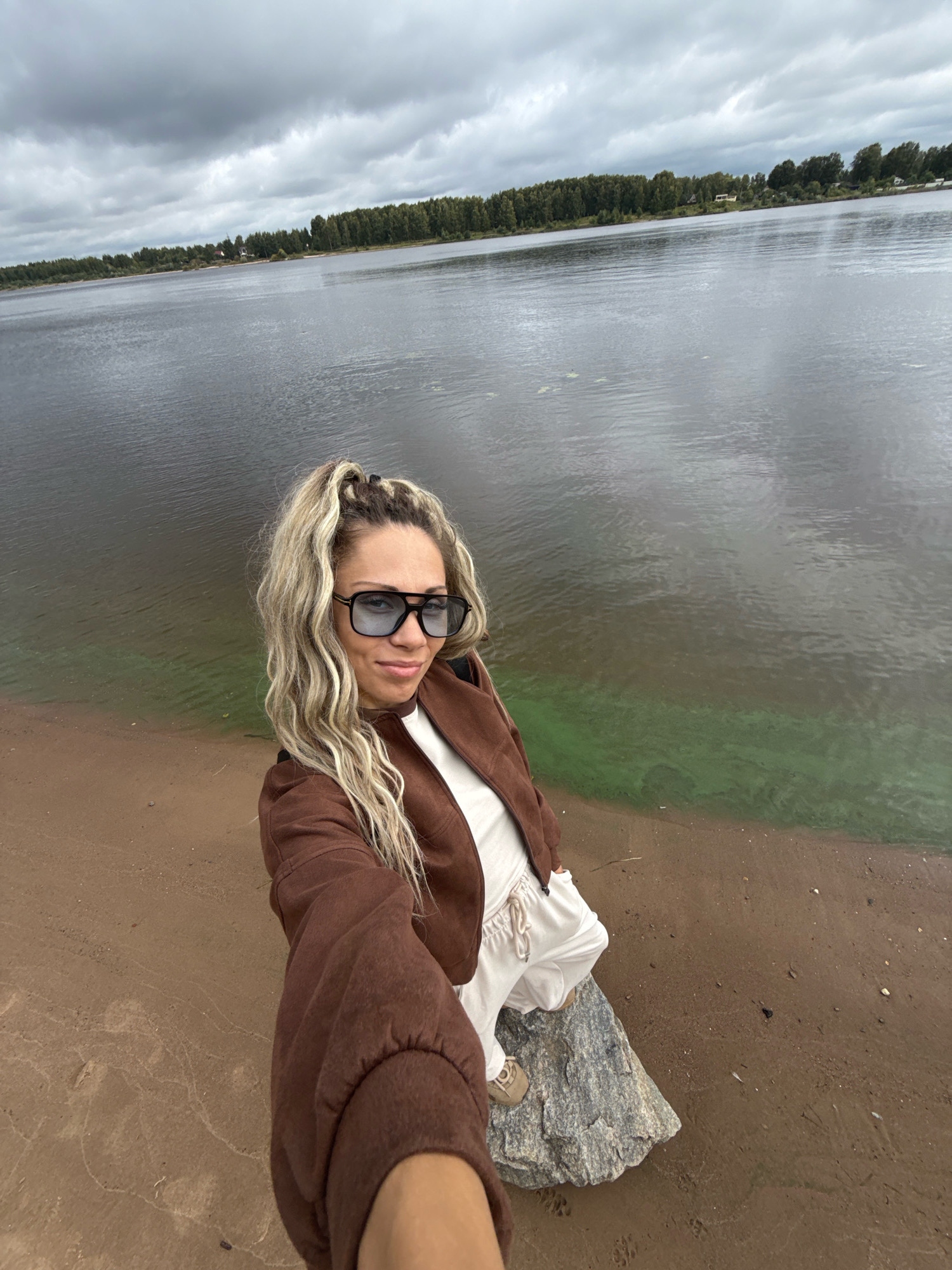 A woman is taking a selfie by a body of water. She is wearing a brown jacket, white top, and white pants. She has blonde hair styled in a ponytail and is wearing sunglasses. She is standing on a rock next to a sandy shore. The water is calm and appears murky with some green algae visible near the shore. In the background, there is a treeline and a cloudy sky. The scene appears to be in Ignatovo, Russia, likely near the Oka River, given the presence of the vast body of water and the treeline consistent with the region.