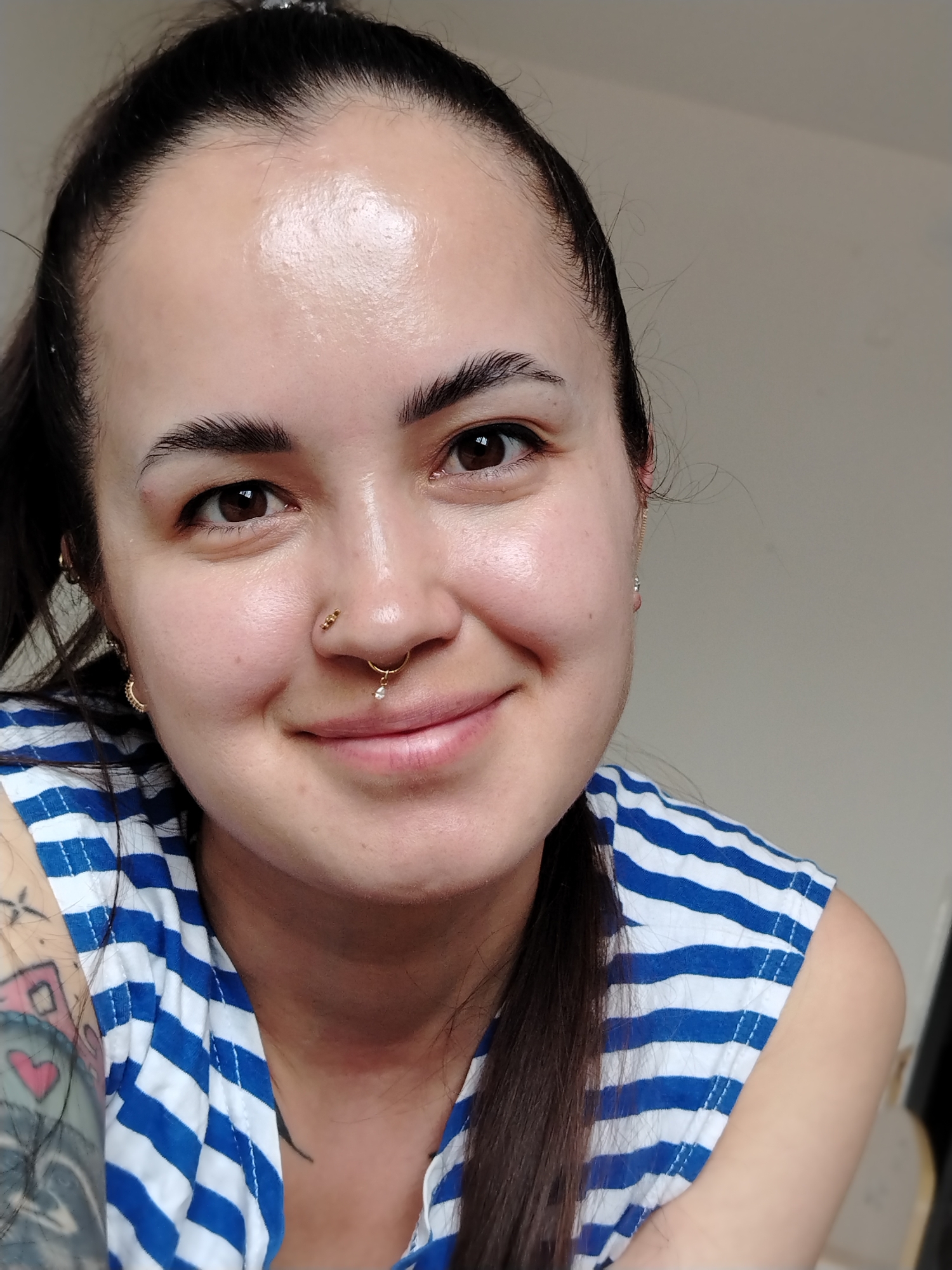 A close-up shot features a light-skinned woman with brown eyes and brown hair pulled back in a ponytail. She is smiling slightly, revealing a hint of her upper teeth. Her nose is adorned with two gold nose rings, one a small stud and the other a septum ring with a dangling gem. She is wearing a blue and white striped sleeveless shirt. Part of a tattooed arm is visible on the left side of the frame. The background is a plain, light-colored wall. The photo seems to be taken indoors, with natural light illuminating the subject's face. Location is unknown.