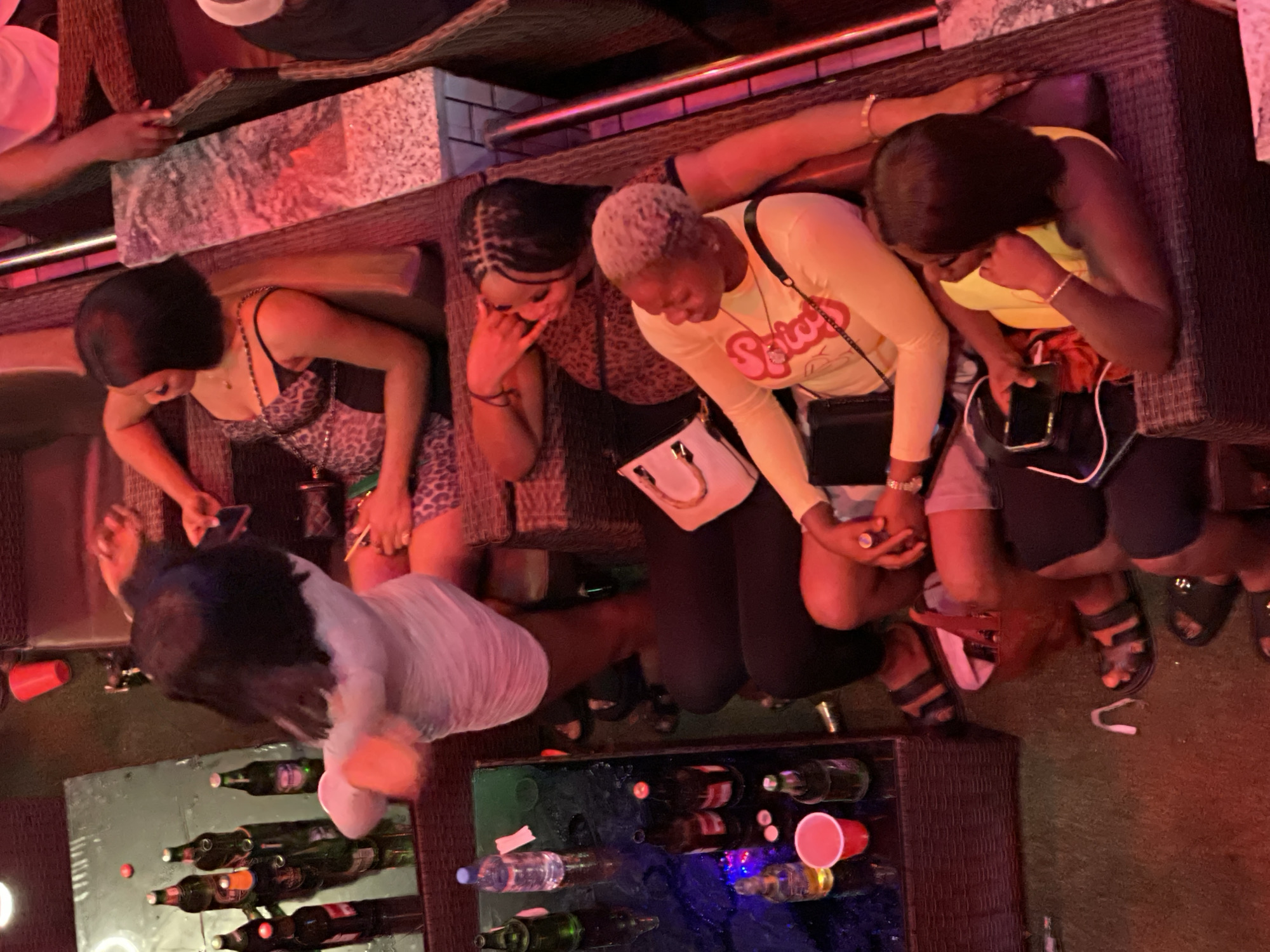 In the city of Unknown, Unknown, an aerial view captures a dimly lit, outdoor lounge area with multiple individuals seated on wicker furniture. A glass table in the foreground is laden with several green glass bottles and red plastic cups. One woman, seen from above, wears a white dress and has dark, straight hair. She appears to be in motion, with her arms outstretched. Another woman, also viewed from above, sports a leopard-print top with spaghetti straps and dark hair. A third woman has short, blonde hair and wears a yellow long-sleeved top. A fourth woman, with dark braided hair, is positioned behind the woman in the yellow top. The scene suggests a casual gathering with drinks, potentially at a bar or club. The lighting is predominantly red and ambient.