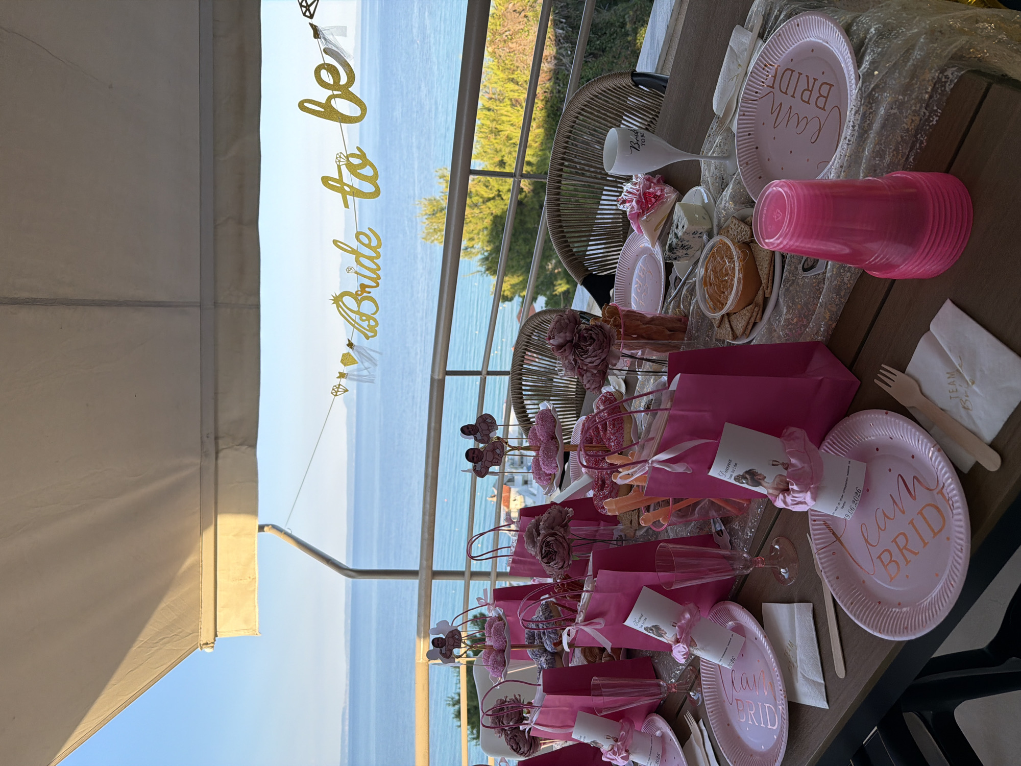 The image depicts a bridal shower setup on an outdoor balcony overlooking the sea, likely in Kassandra Municipal Unit, Greece, given the clear blue waters and coastal vegetation. A table is laden with pink paper plates, pink paper cups, pink gift bags, and decorative items. A "Bride to Be" banner is hung across the railing. Some of the plates have "Team Bride" written on them in rose gold lettering, and some contain personalized cards with an image of a bride. Food items, including donuts, cheese, crackers, and a dip, are also visible on the table. The overall atmosphere suggests a festive occasion in a scenic location.