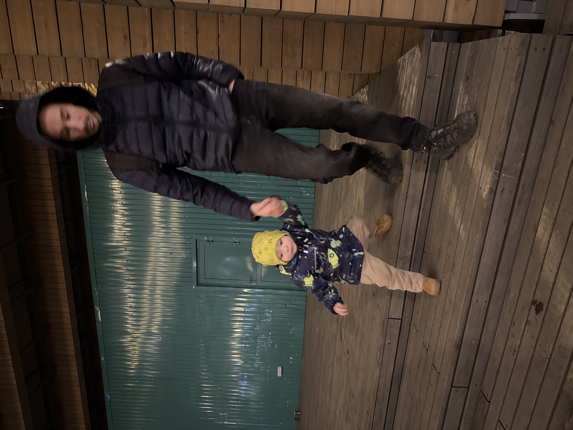 A man and a young child are standing outside on a wooden surface, possibly a deck or elevated platform. The man wears a dark puffer jacket with the hood up and dark pants. The child is dressed in a patterned jacket, light-colored pants, and a yellow hat. They are situated near a green wall with a door. The location is in Izhevsk, Russia.