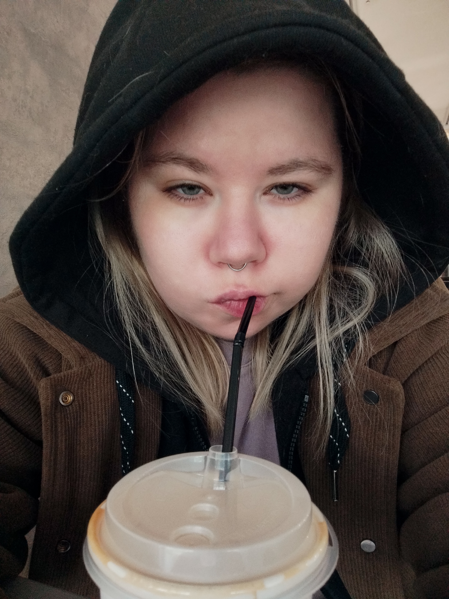A young woman is captured in a close-up, frontal shot, wearing a black hooded sweatshirt over a brown corduroy jacket. She has blonde hair and a septum piercing, and her lips are pursed as she sips from a clear plastic cup with a brown beverage. The background is a textured, light brown wall. The woman's expression appears neutral or perhaps slightly bored. The lighting is soft and diffused.