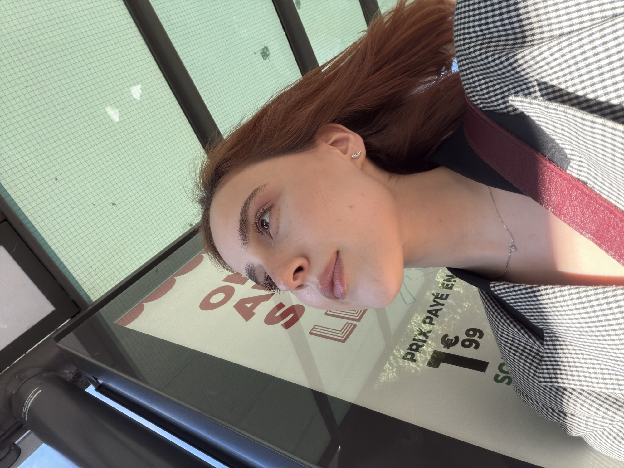 In a low-angle shot, a young woman with auburn hair looks up towards the right. She is wearing a black and white checkered blazer over a red strap. A delicate silver necklace with a small pendant adorns her neck. She has on small, sparkly earrings. In the background, a sign with red lettering is visible, partially obscured by the woman. The text on the sign includes "PRIX PAYÉ EN" and "1€99". The environment suggests an outdoor setting, possibly a bus stop, with a translucent green roof overhead supported by a dark grey pole. Sunlight filters through the roof, casting dappled shadows. The image appears to have been taken in the city of Strasbourg, France.