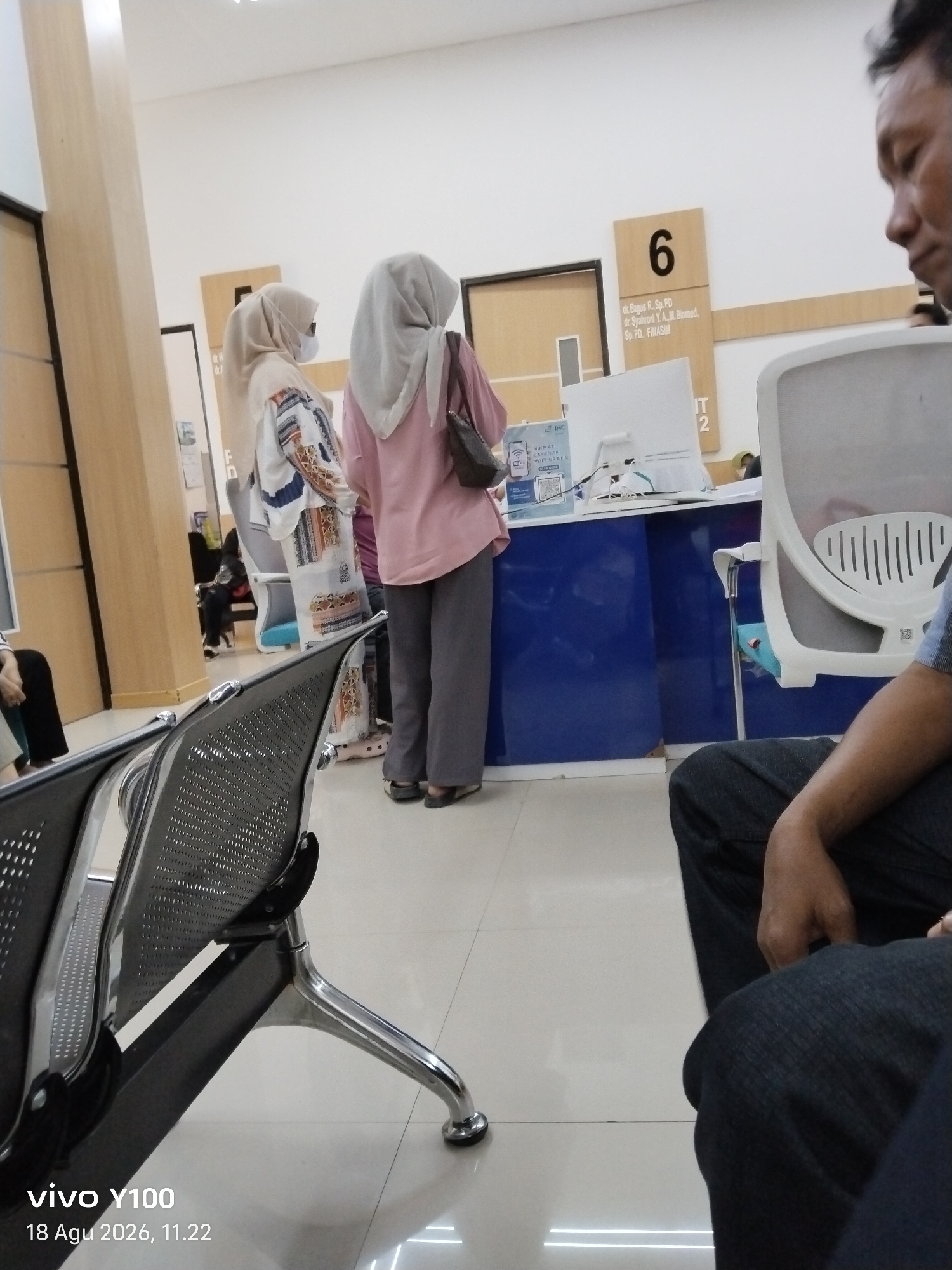 The image depicts the interior of what appears to be a waiting area in a medical facility, possibly a clinic or hospital, in Patokan, Indonesia. In the foreground, a metal waiting chair with a perforated seat is visible. To the left, a man in a blue shirt and dark jeans is seated, with his right hand resting on his lap. In the midground, two women wearing hijabs are standing and facing away from the camera. The woman on the left wears a white and patterned traditional outfit, and the woman on the right wears a pink top and grey pants. Behind them, a blue reception desk is situated, with a computer monitor and a sign that reads "NIKMATI LAYANAN WIFI GRATIS" (Enjoy Free WiFi Service). A sign with the number "6" and doctor's names is mounted on the wall above the desk. The environment is brightly lit, with tiled flooring. The timestamp at the bottom left corner of the image indicates "vivo Y100 18 Agu 2026, 11.22".