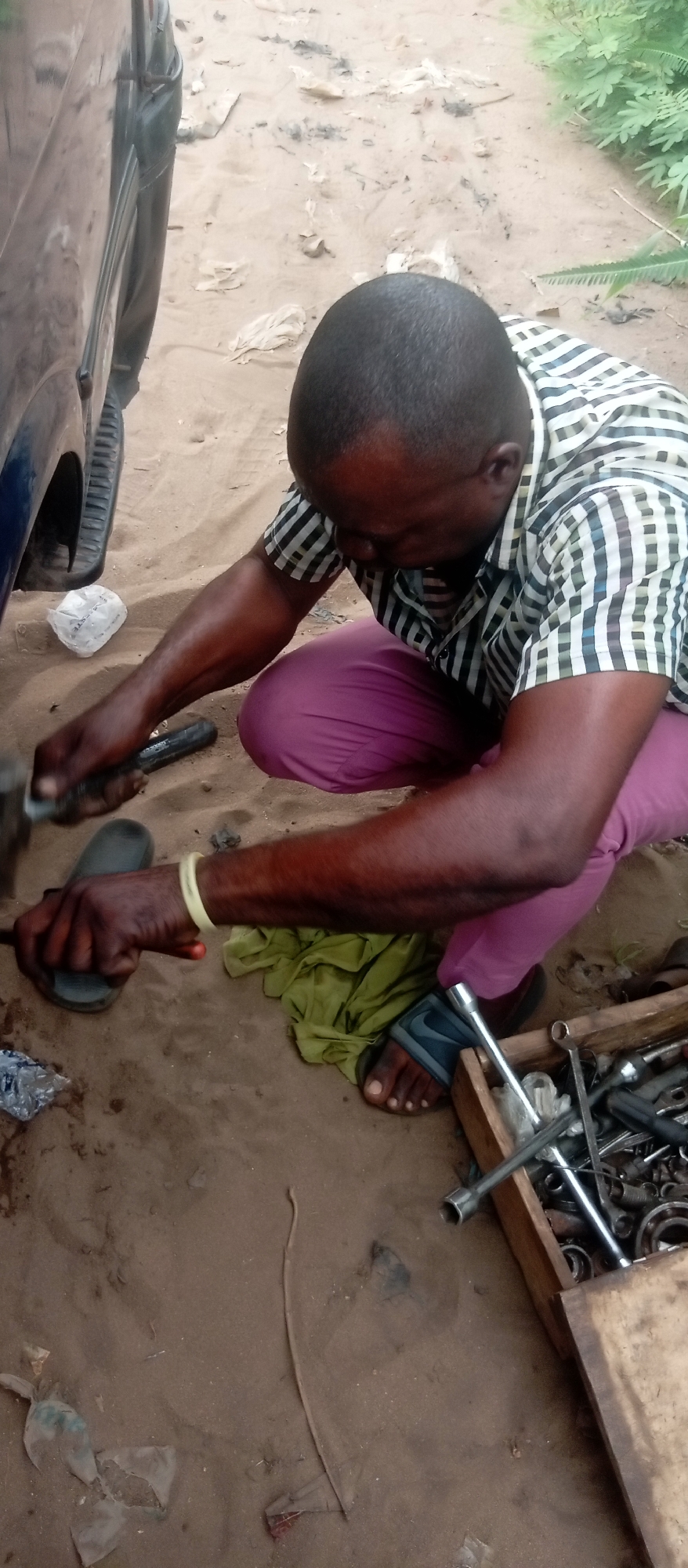 A man is crouched down working on a car in an outdoor environment. He is wearing a short-sleeved, checkered shirt and pink pants, with sandals on his feet. He is holding tools in his hands and appears to be repairing something on the car, which is visible on the left side of the frame. Various tools are scattered on the ground and in a wooden box next to him. The ground is sandy, with scattered debris and a piece of green cloth. The location is in Ogbunike, Nigeria, as this information was pre-provided. The scene suggests a makeshift auto repair setup in a relatively unstructured environment.