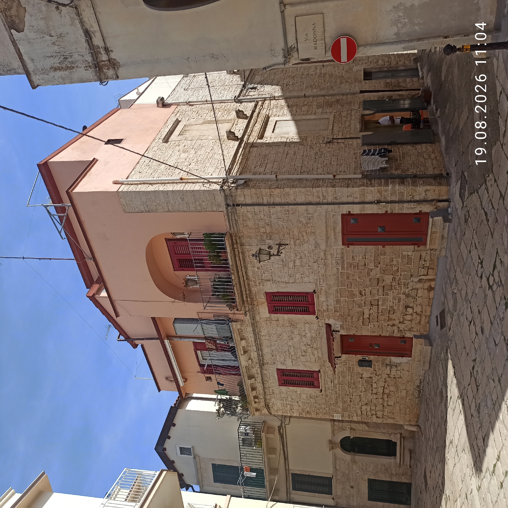 The image shows a narrow street in Ruvo di Puglia, Italy, paved with stone, with buildings on either side. The main building in the foreground is made of light-colored stone blocks and has red doors and window shutters. The upper part of this building is painted pink and features a balcony with a metal railing. Adjacent to it, on the left, is another building with white walls and dark green shutters. On the right, a sign reads "VIA MADONNA" above a red and white circular "no entry" sign. Laundry is hanging from a balcony of the pink building. A person in a white shirt is visible through an open doorway of a building on the right side of the street, and an orange chair is placed near the entrance. The sky is clear and blue. The timestamp "19.08.2026 11:04" is overlaid at the bottom right of the image.