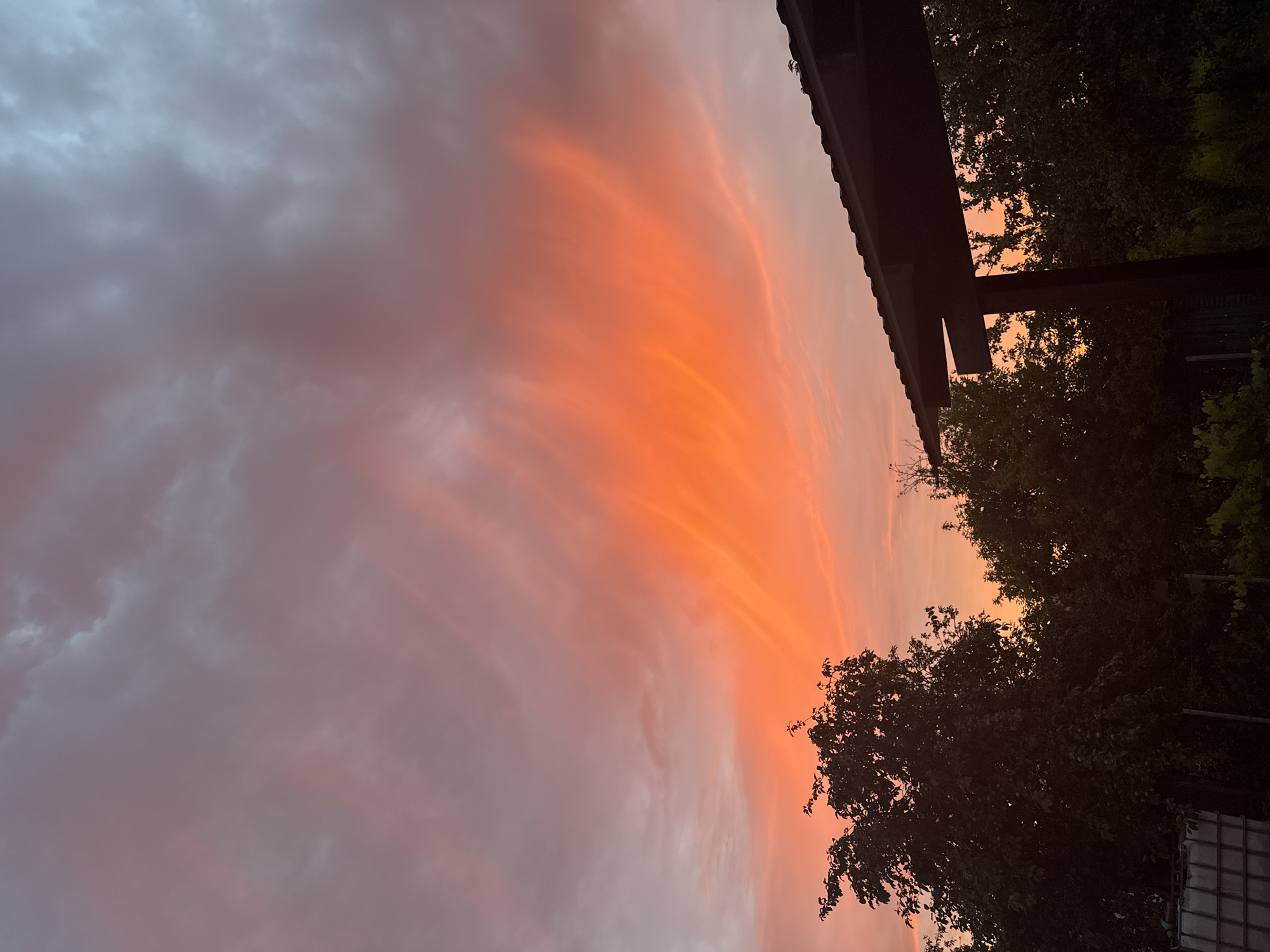 The image depicts a vibrant sunset over a rural or suburban landscape. The sky is dominated by dramatic clouds, with a brilliant orange and red hue concentrated in the center, gradually fading into softer pinks and purples towards the edges. Wispy grey clouds are interspersed throughout, contrasting with the intense colors of the sunset. Silhouetted against the colorful sky are the dark shapes of trees, their foliage dense and full. In the foreground, on the right side of the image, a dark, angular structure, possibly a roof or awning, partially obstructs the view of the sky and trees. On the lower left, a portion of a large white industrial-looking tank or container is visible, along with some fencing and foliage. The scene is tranquil, capturing the ephemeral beauty of a sunset. No people or specific landmarks that would indicate a precise city or urban environment are present.