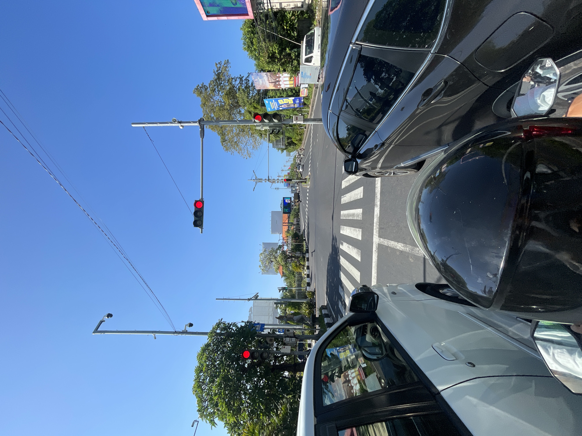 The image depicts a traffic scene at an intersection in Kuta Selatan, Indonesia, under a clear blue sky. The perspective is from the vantage point of a motorcyclist, as indicated by the handlebars and rearview mirror in the foreground. Two cars, a white one on the left and a dark-colored SUV on the right, are stopped in traffic lanes, aligned with white pedestrian crossing lines on the asphalt.
Several traffic lights are visible. The closest traffic light on the left is red, as is another on the right. Further down the road, two more traffic lights are also illuminated red. Traffic poles with cameras and electrical wires are present throughout the scene. Trees with lush green foliage border the road, and some billboards are visible in the background. In the distance, other vehicles, including cars and motorcycles, are also stopped or moving slowly. The overall impression is of a typical urban intersection experiencing moderate traffic congestion during daylight hours.