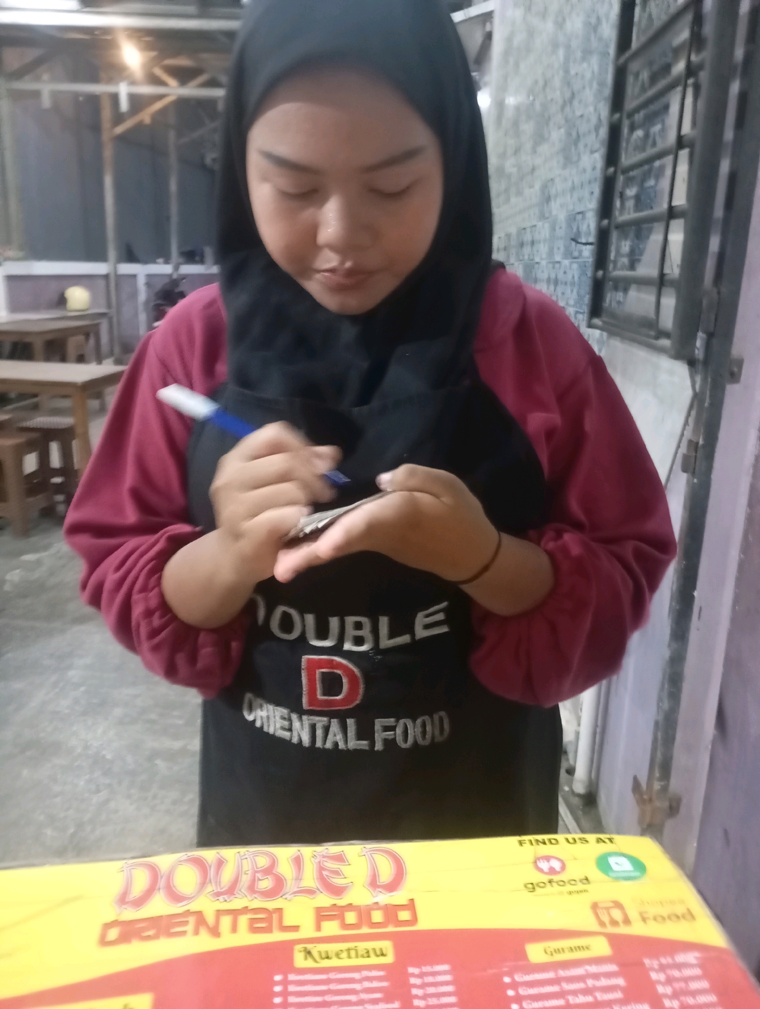 A young person wearing a black hijab and a maroon sweater over a black apron with the "DOUBLE D ORIENTAL FOOD" logo is standing and writing on a small stack of papers with a blue and white pen. They are holding the papers in their left hand, which is positioned over their stomach. A thin black bracelet is visible on their right wrist. The background shows an outdoor or semi-outdoor food stall setting. To the left, there are wooden tables and chairs. To the right, a tiled wall with metal bars is visible. In the foreground, a yellow sign with red and white text, also displaying "DOUBLE D ORIENTAL FOOD," along with logos for "gofood" and "Shopee Food," is partially visible. The scene is dimly lit, suggesting it is either evening or an indoor space with artificial lighting. The location is identified as Babelan, Indonesia.