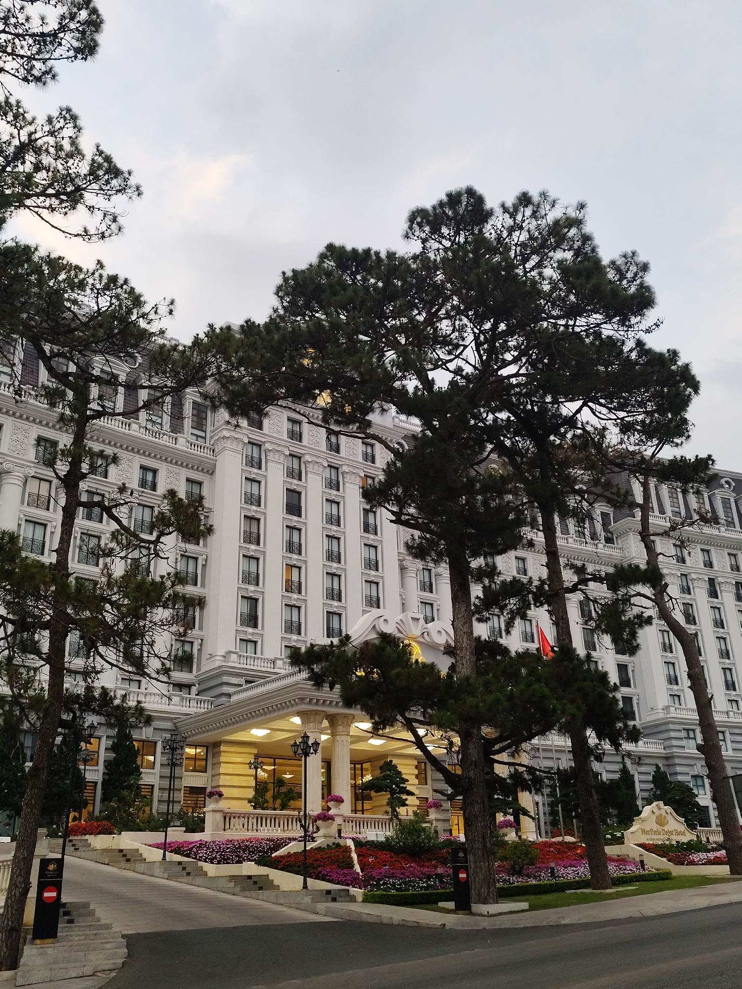 The image depicts the Mercure Dalat Resort, a prominent landmark in the city of Da Lat, Vietnam. The multi-story, white building exhibits classical French architectural details. Large pine trees partially obscure the view of the building, framing the entrance. A meticulously maintained flower garden in vibrant colors is located in front of the building. A Vietnamese flag is visible amongst the trees. The sky is overcast, suggesting either early morning or late afternoon. A paved road and walkways lead up to the entrance of the resort. A sign with the hotel's name stands in front of the garden. No people are clearly visible in the image.