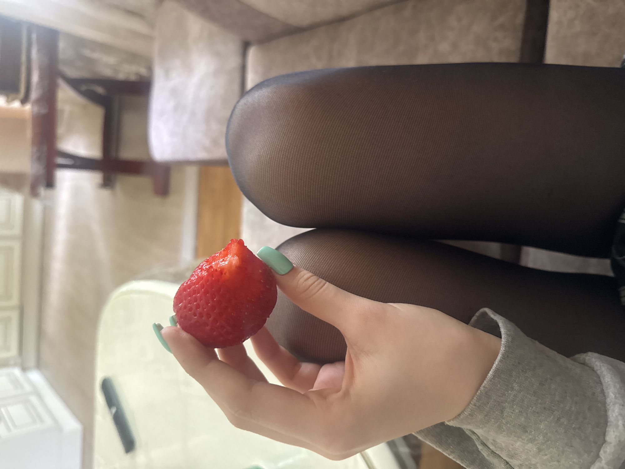A medium, close-up shot shows a person holding a strawberry. The person has a light complexion and is wearing a gray long-sleeved shirt. They are holding the strawberry with their left hand, and their nails are painted a light green color. The strawberry is red and has had a bite taken out of it.
The person is wearing black tights. The background includes a portion of a beige couch or chair, and a blurred view of what appears to be a kitchen or dining area with white cabinets or walls. There is also a blurred glimpse of wooden furniture.
The photograph does not contain information that could confirm a location in Saint Petersburg, Russia.