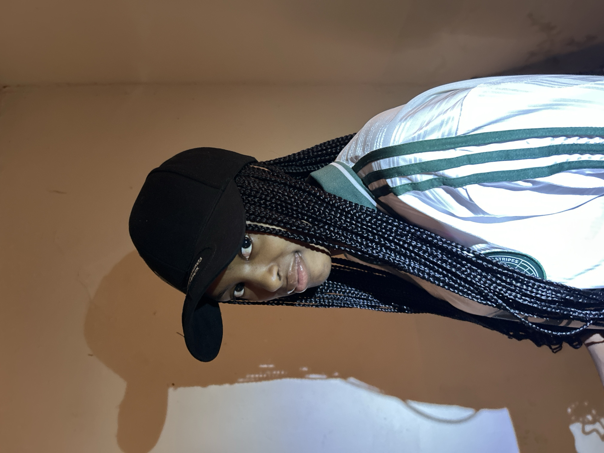 A young Black woman with braided hair and a black baseball cap is photographed in what appears to be a room in Jos, Nigeria. She is wearing a white and green athletic jacket with three green stripes down the sleeves and across the chest. Her gaze is directed slightly to the left of the camera. A large, indistinct shadow is cast on the wall behind her, to the left of the frame. The lighting in the room is dim, with a warm tone on the wall.