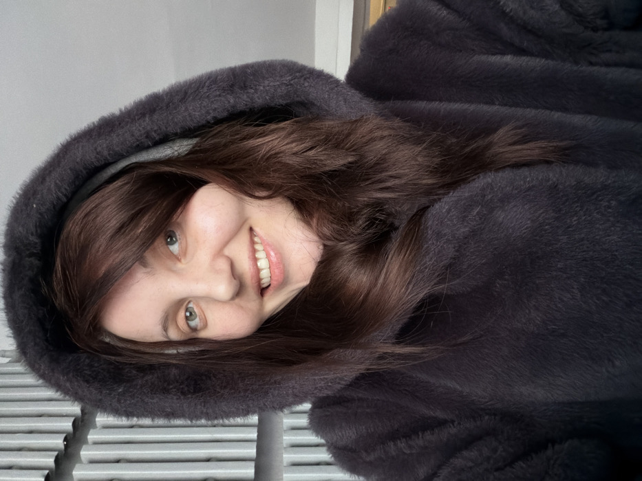 A woman with fair skin, green eyes, and long brown hair smiles while wearing a dark gray hooded fur coat. She is tilted slightly to the side, with her face illuminated and teeth visible. The hood frames her face, and soft, gray fur surrounds her. Metal bars are visible in the lower part of the frame, possibly a railing or grate.