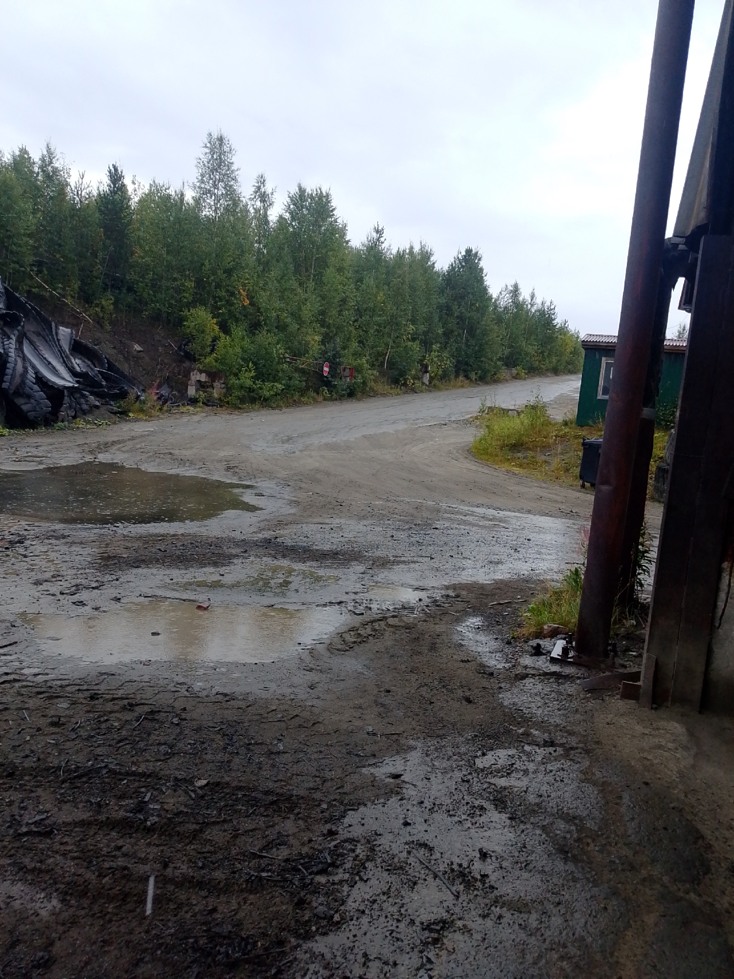 An outdoor, medium shot captures a muddy, rural road curving through a dense forest under an overcast sky. The road, covered in puddles and debris, is flanked by a steep, tree-covered embankment on the left and industrial structures on the right. A large pile of black, burnt material, possibly tires, is visible on the embankment. To the right, a dark green building with a white-framed window stands beside a rusted metal pipe. The foreground is dominated by dark, wet mud with tire tracks and scattered small objects.