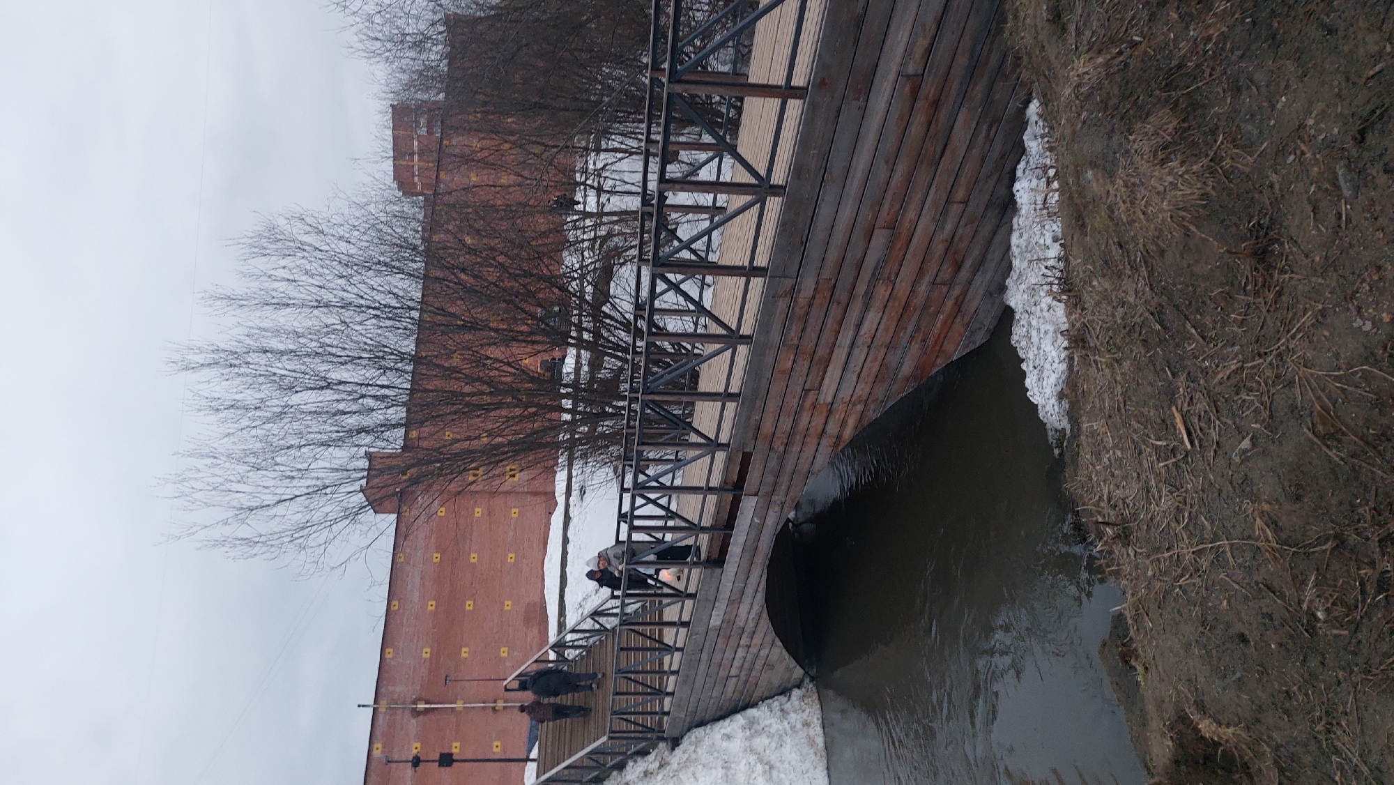 The image depicts a wintry scene with a wooden bridge spanning a body of water. The bridge has a metal railing and a pedestrian walkway. On the left side of the image, a tall brick building with a grid of yellow squares is visible. Bare trees line the bridge, obscuring some of the building. People are walking on the bridge. Snow is present on the banks of the water, and the ground appears muddy. The sky is overcast. This is the embankment of the Ob River in Novosibirsk, Russia.