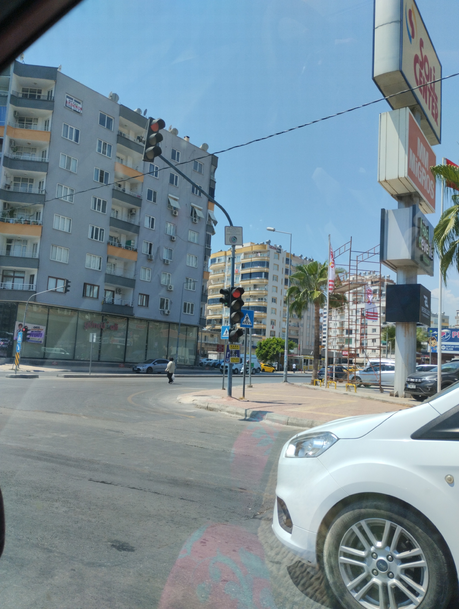 The image captures a street scene in Mezitli, Turkey, under a clear blue sky. A multi-story apartment building with grey and orange accents dominates the left side of the frame, featuring numerous windows and balconies. In front of this building, a street intersection is visible with a traffic light displaying a red signal. A pedestrian is crossing the street.
To the right of the apartment building, a tall sign with "ÇOLAK CENTER" is illuminated, and below it, another sign reads "MIM MIGROS". A large construction scaffold stands near the "MIM MIGROS" sign, adorned with red and white striped flags. Several palm trees are scattered throughout the scene, contributing to the urban environment.
The foreground is partially obscured by the front of a white vehicle, showing its wheel and part of the fender. The road surface is asphalt, with some dark patches and tire marks. In the background, other buildings and vehicles can be seen, including a yellow taxi. The overall impression is a bustling urban intersection on a sunny day.