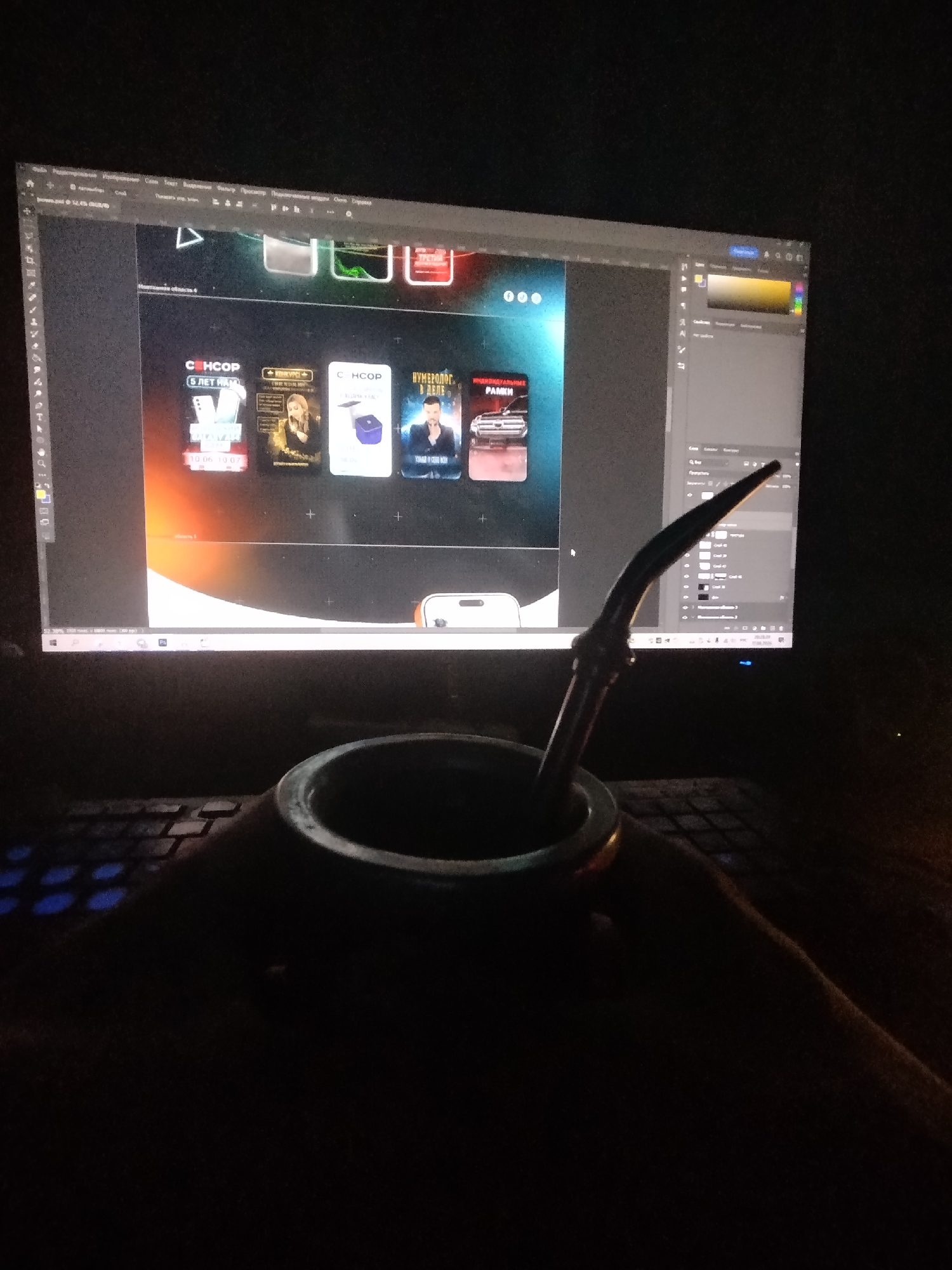 A dimly lit indoor scene features a computer monitor displaying a graphic design interface, likely Adobe Photoshop, judging by the tools and panels visible. Several digital mockups of book covers or app icons are arranged horizontally on the screen. In the foreground, a person's hands are holding a dark, rustic-looking mate gourd with a metal bombilla, a traditional South American sipping straw. The mate and bombilla are positioned over a keyboard with some keys illuminated by a blue backlight. The overall ambiance is that of someone working or creating late at night, possibly enjoying a beverage while engaged in digital design work. The location is not discernible from the image.