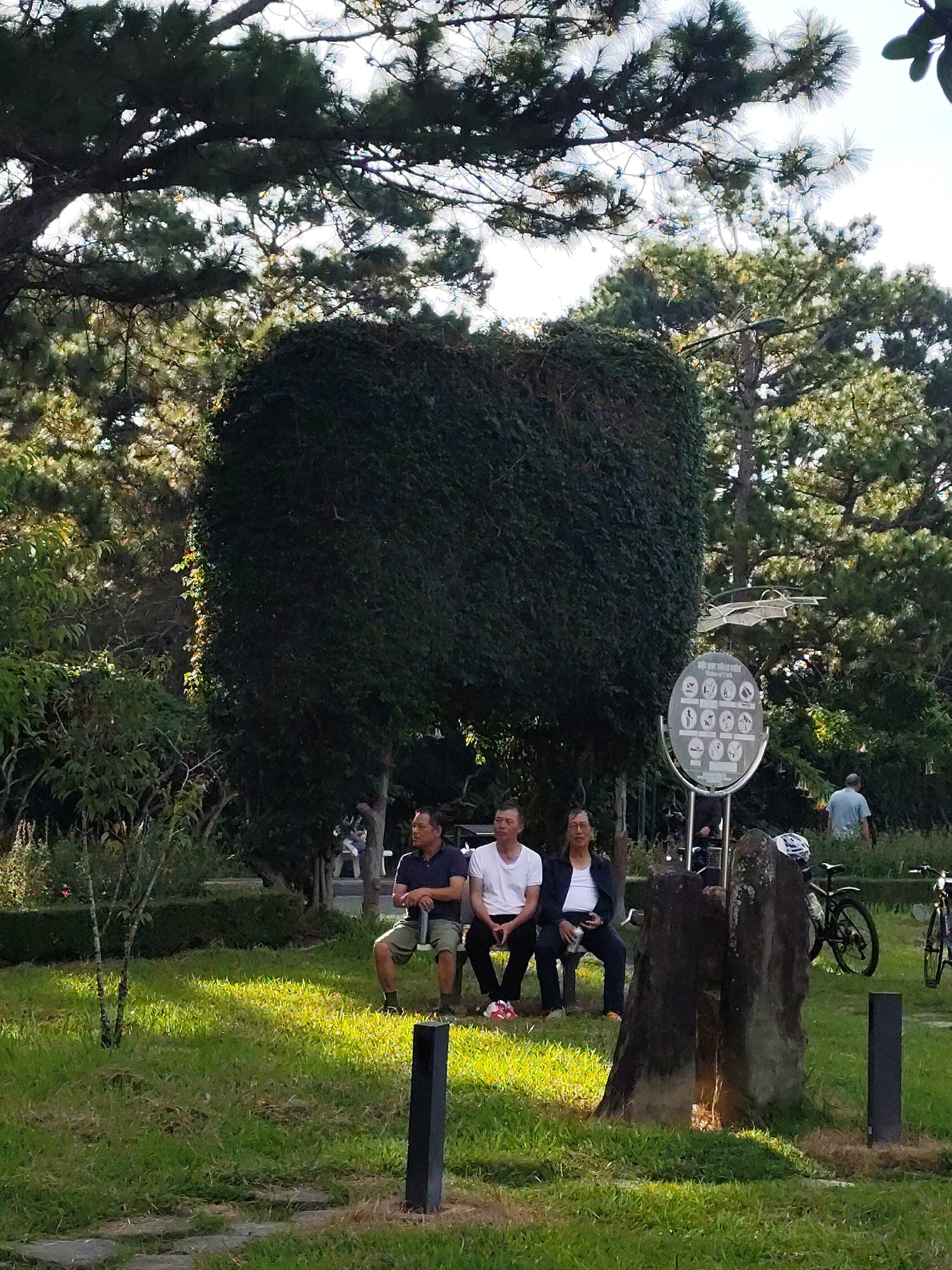 The image depicts a scene in a park in Xuân Hương - Đà Lạt, Vietnam, characterized by lush greenery and a bright, sunny atmosphere. In the foreground, three men are seated on a bench. The man on the left wears a white t-shirt and black pants, with red and white sneakers. The middle man is dressed in a white undershirt, a dark suit jacket, and dark pants. He holds a silver can in his right hand. The man on the right is in a dark polo shirt and khaki shorts. Behind them, a large, dark green, square-shaped hedge dominates the center of the frame. To the right of the hedge, a circular sign with multiple icons is mounted on a metal stand. Further to the right, two bicycles are parked, one black and one dark blue. A man in a light blue t-shirt walks away from the camera in the background. The ground is covered in green grass, with some stone pathways visible. Tall trees with dense foliage form the backdrop, their branches reaching towards the sky, filtering the sunlight. The overall impression is one of a peaceful, outdoor gathering space.