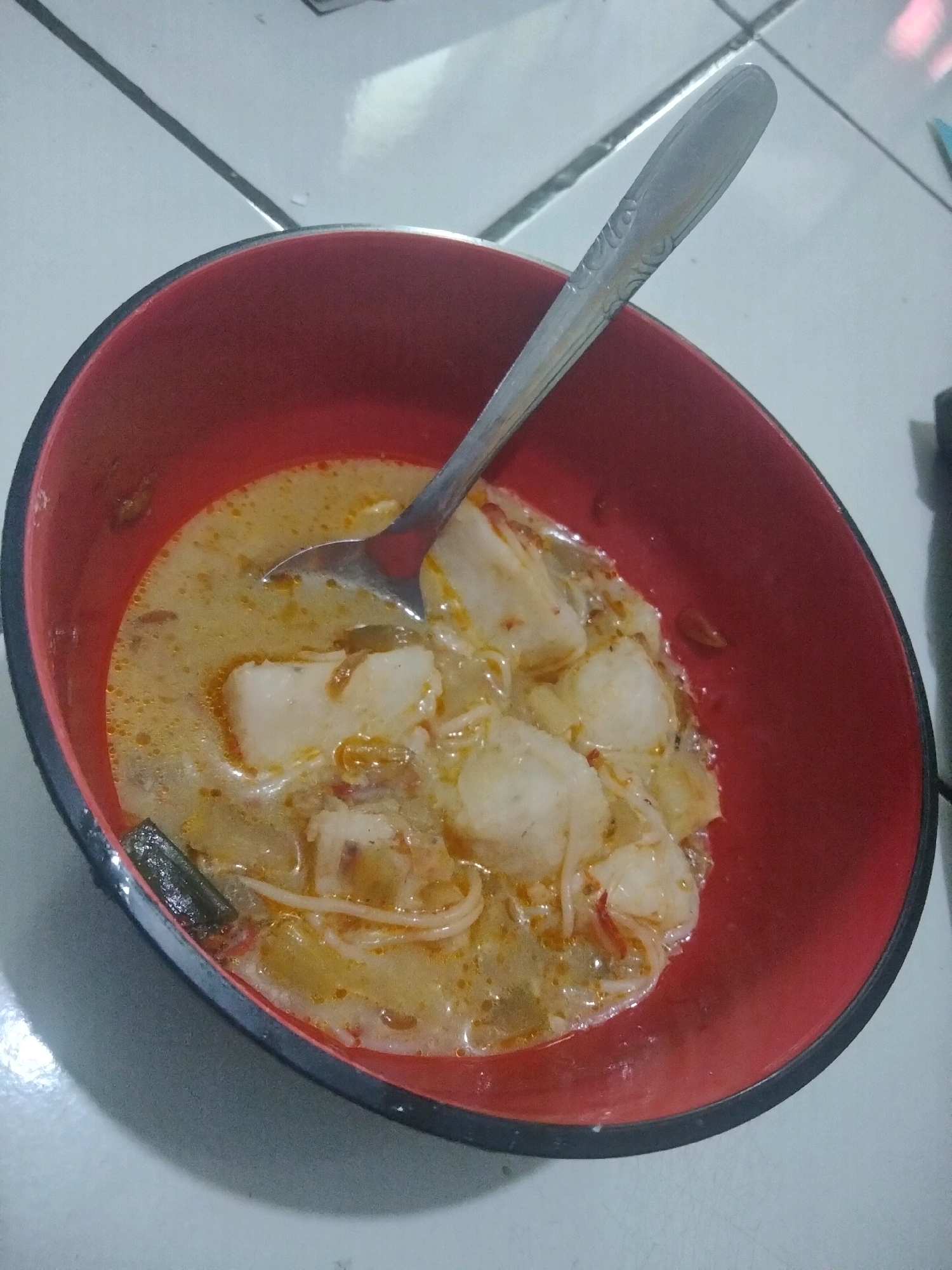 A red and black bowl filled with a creamy, yellowish broth containing white meatballs, noodles, and what appears to be chunks of white protein, is positioned on a tiled surface. A metal spoon with an embossed pattern rests in the bowl, partially submerged in the broth. The tiled surface has visible grout lines and appears to be an indoor floor. The overall scene suggests a meal being served or consumed. The provided information suggests this image was taken in Suka Sari, Indonesia.
