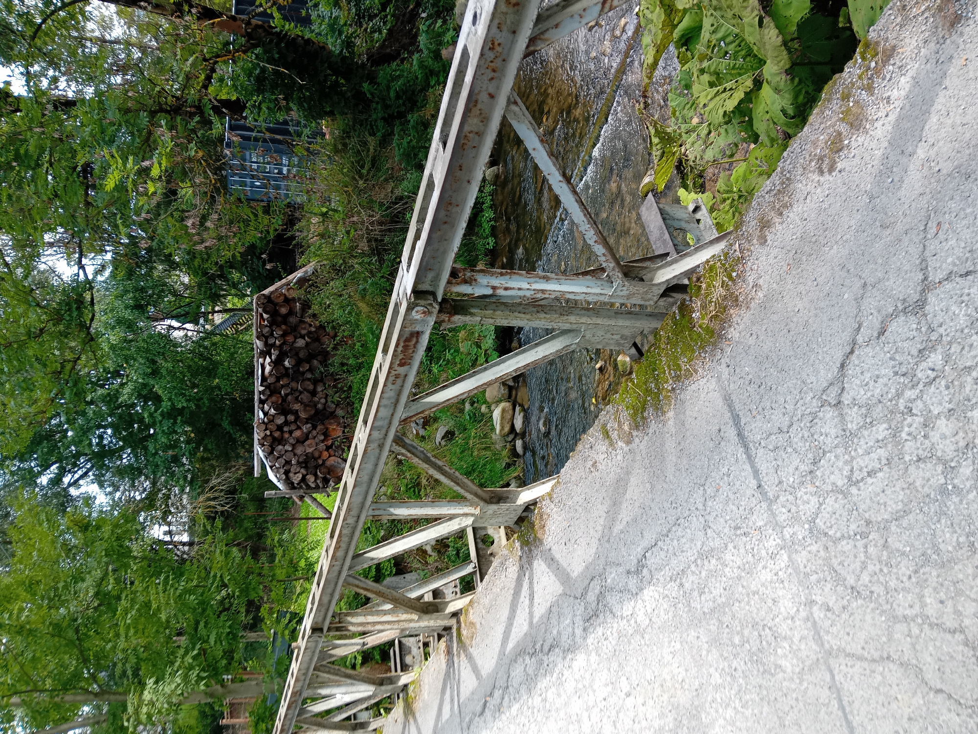 The image shows a rustic metal bridge crossing a shallow, rocky stream in Zuberec, Slovakia. The bridge has a worn, gray metal railing with visible rust and is supported by a lattice of metal beams. The road surface on the bridge is cracked asphalt, with patches of moss. To the right of the bridge, a stack of freshly cut logs is visible, along with dense green foliage and trees. In the background, a large blue shipping container can be seen amongst the trees. The stream bed is made up of stones and pebbles, with clear water flowing over them. The environment appears to be rural and somewhat unkempt.