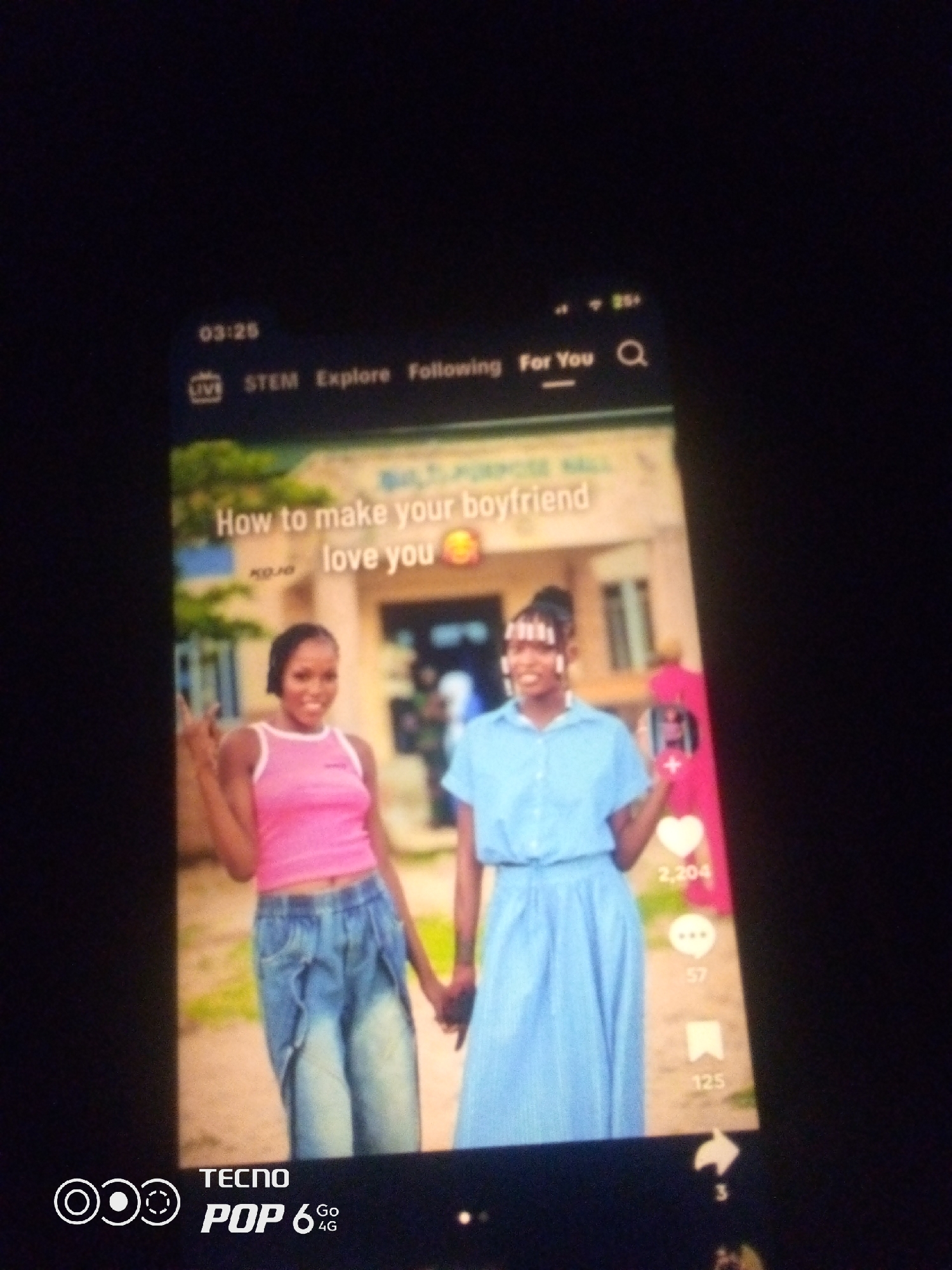 The image shows a smartphone screen displaying a TikTok video. The video features two young women standing in front of a building labeled "MULTI PURPOSE HALL". The woman on the left wears a pink tank top and baggy blue jeans, while the woman on the right wears a light blue collared shirt and a blue skirt. They are holding hands. Text on the screen reads "How to make your boyfriend love you". The location in the city of Howayi, Nigeria is not identifiable from the provided image. The smartphone also shows social media interface elements like likes, comments, and shares.