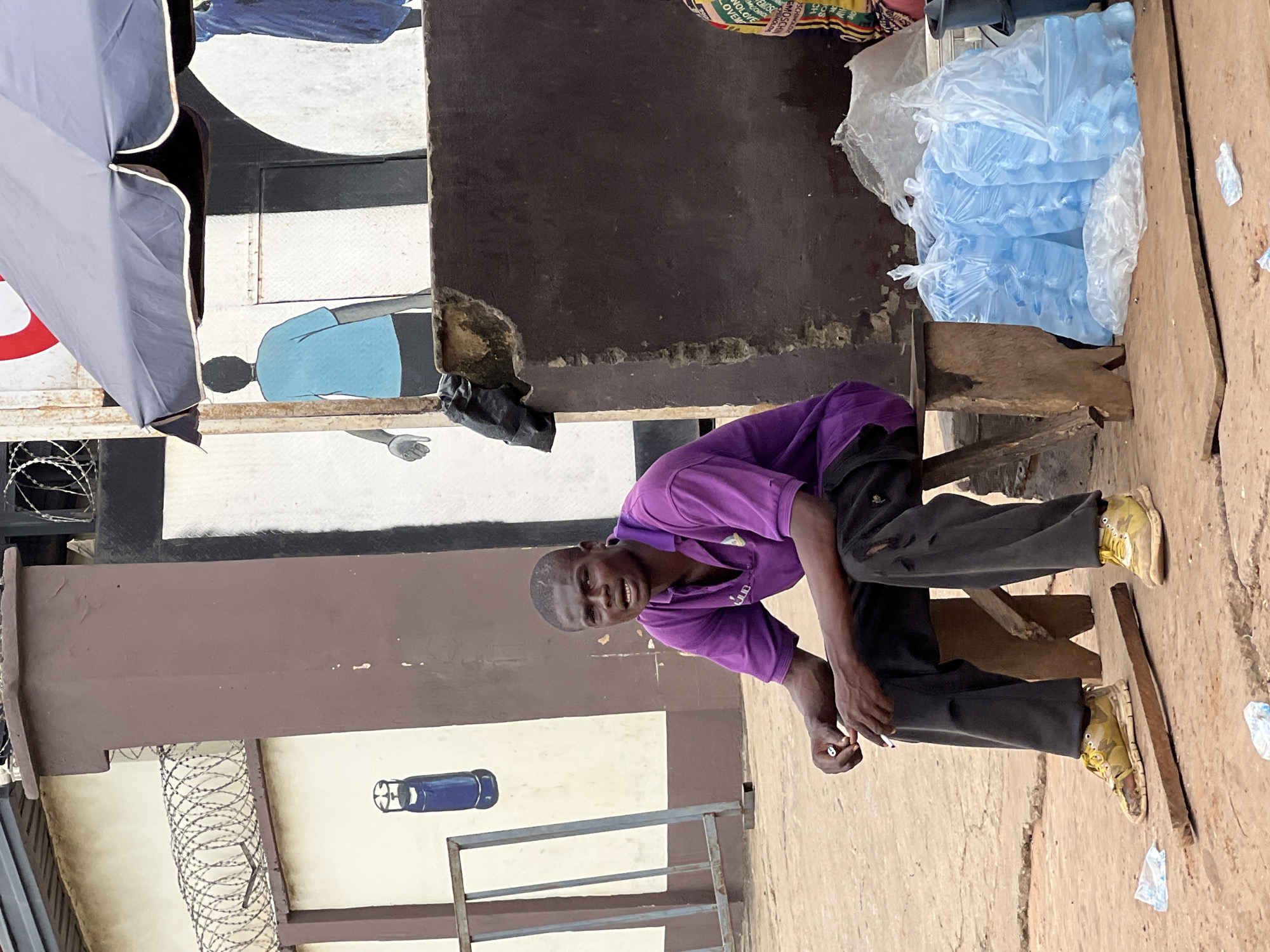 A Black man sits on a wooden stool in Nkpor, Nigeria, holding a cigarette. He wears a purple polo shirt and ripped dark pants, with yellow sneakers. The man is positioned in front of a brown, weathered wall and a mural depicting figures. Behind him, a dark umbrella shades a stall. In front of the man, several plastic bags filled with blue liquid, likely water sachets, are stacked. The ground is dusty and appears to be a street or marketplace.