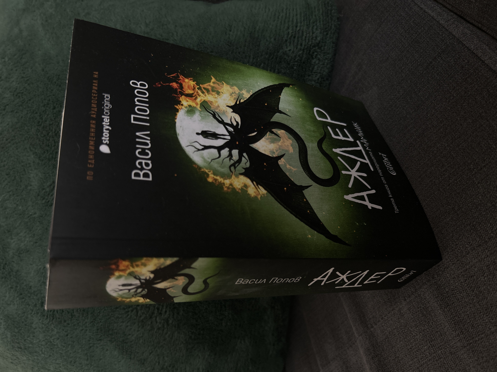 The image shows a paperback book titled "АЖДЕР" by Васил Попов, translated as "Azhder" by Vasil Popov. The book is dark green with black accents and features an illustration of a winged dragon-like creature in front of a full moon amidst flames. The cover also has text that says "ПО ЕДНОИМЕННИЯ АУДИОСЕРИАЛ НА storytel original", "Трета книга от поредицата МАМНИК Glibri", translated as "Based on the audio series of Storytel Original", "Third book in the series Glibri". The book is lying on a grey textured surface, possibly a sofa or blanket. It is positioned at an angle. No specific city is evident from the image.