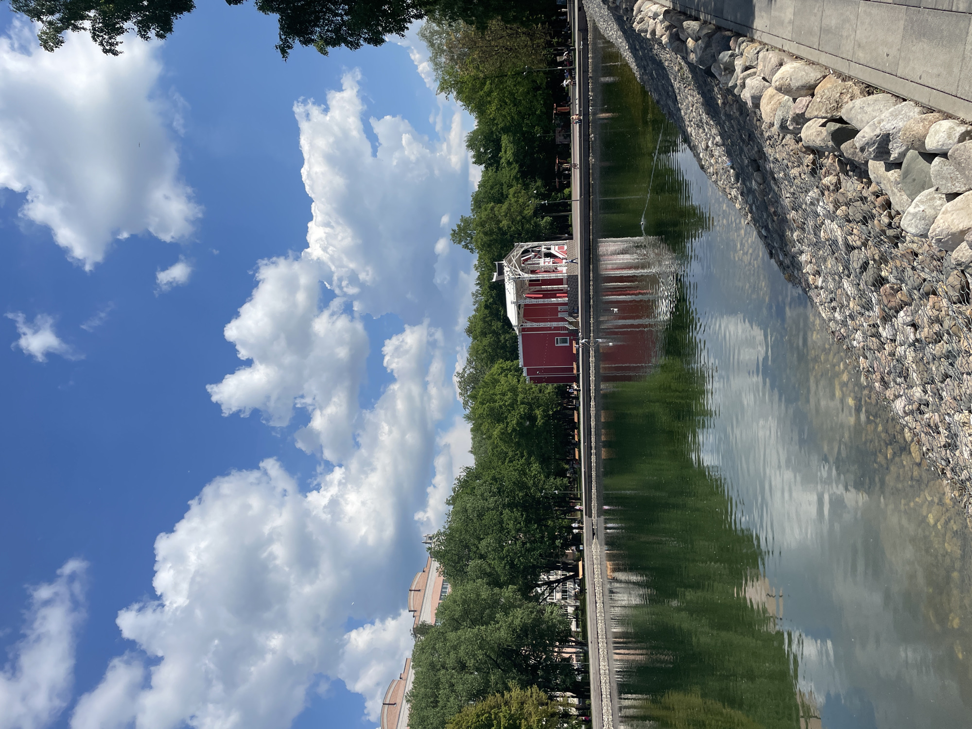 The image depicts a serene park scene with a tranquil pond in the foreground, reflecting the sky and surrounding greenery. A bright red building with white accents, possibly a pavilion or small theater, stands on the far bank of the pond, nestled amidst lush green trees. In the background, a multi-story brick building is visible, suggesting an urban setting. The sky is a vibrant blue, dotted with fluffy white clouds, indicating a pleasant day. The pond's edge is lined with a pathway and a decorative embankment of large stones. The overall impression is one of a peaceful urban oasis, likely a public park in Vologda, Russia, offering a space for recreation and relaxation.