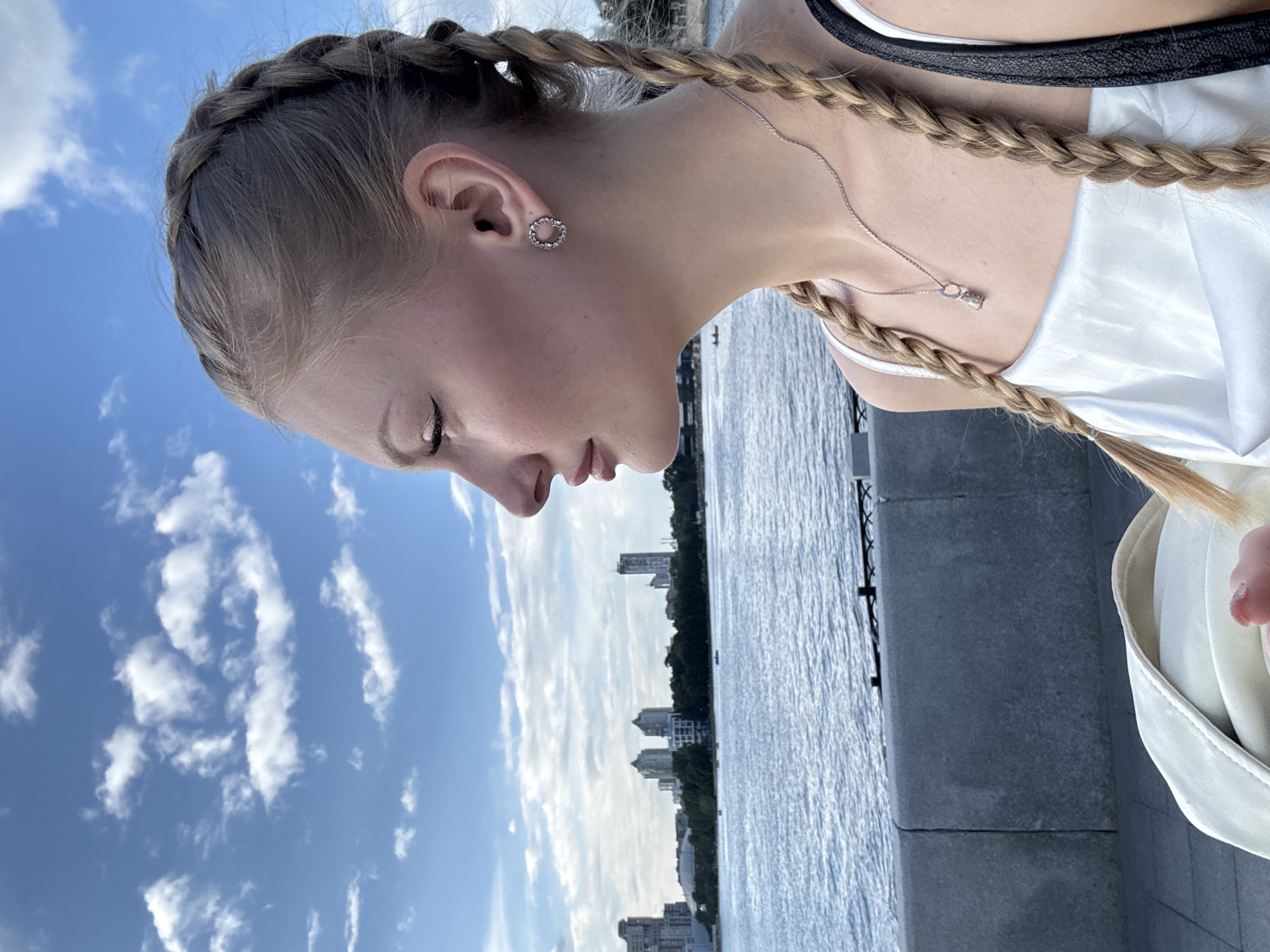 A young woman with blonde hair in two braids is seen from the shoulders up, looking to the left. She is wearing a white dress, a silver necklace with a small pendant, and a circular earring with small stones. The background shows a body of water, likely the Iset River, with a city skyline in the distance, characteristic of Yekaterinburg, Russia. The sky is blue with scattered white clouds, and the lighting suggests it is daytime.