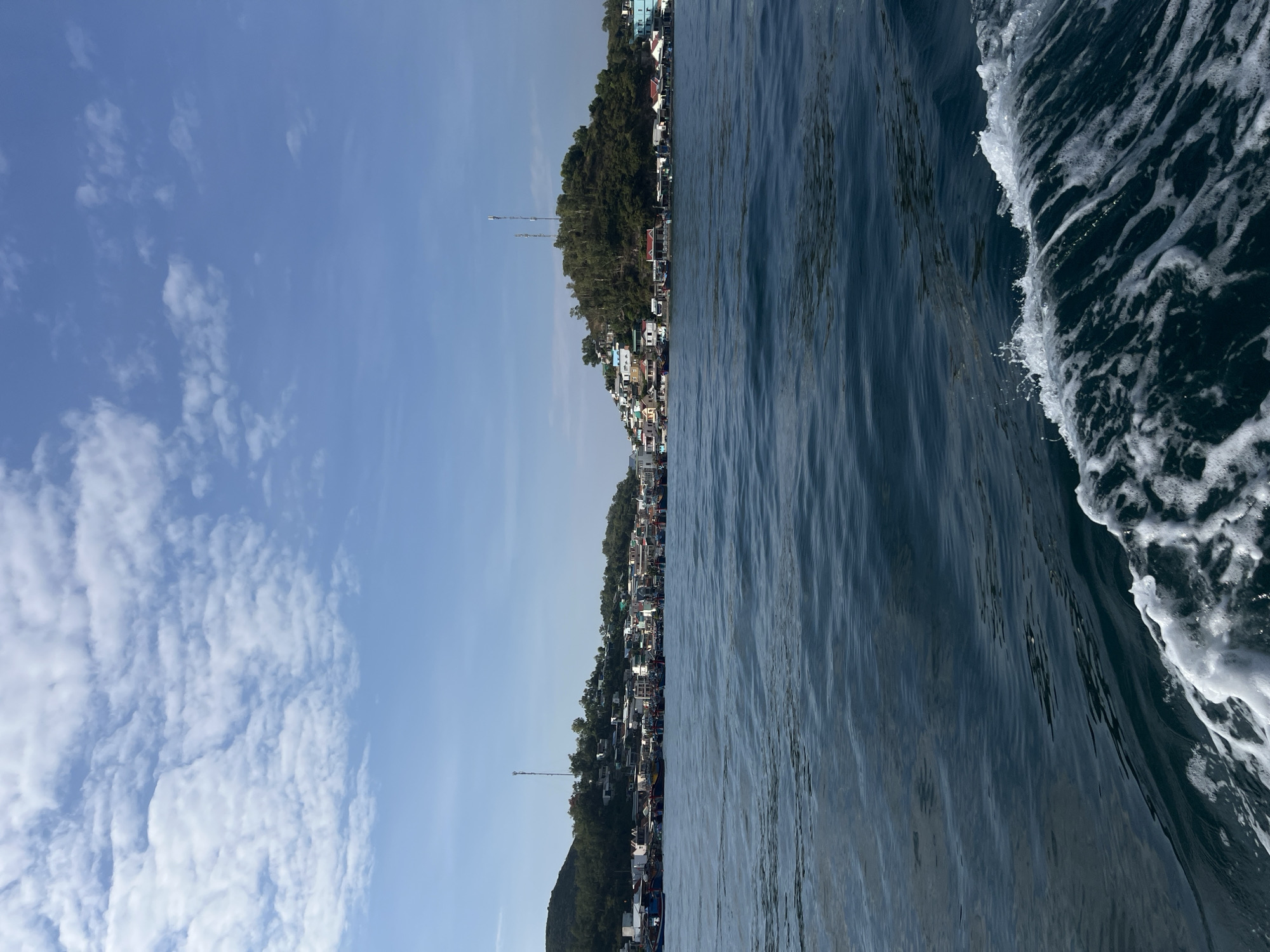 The image depicts a view from a boat moving along the coastline of Nam Nha Trang Ward, Vietnam. On the left side of the image, a clear blue sky with scattered white clouds is visible. Adjacent to the sky, the land is densely populated with buildings, interspersed with trees and vegetation, extending along the coastal strip. Two tall towers or poles can be seen rising above the buildings. To the right of the coastline, dark blue water fills a significant portion of the frame. The wake of the boat is prominently visible as a band of white foam and churning water against the darker water, indicating movement through the water.