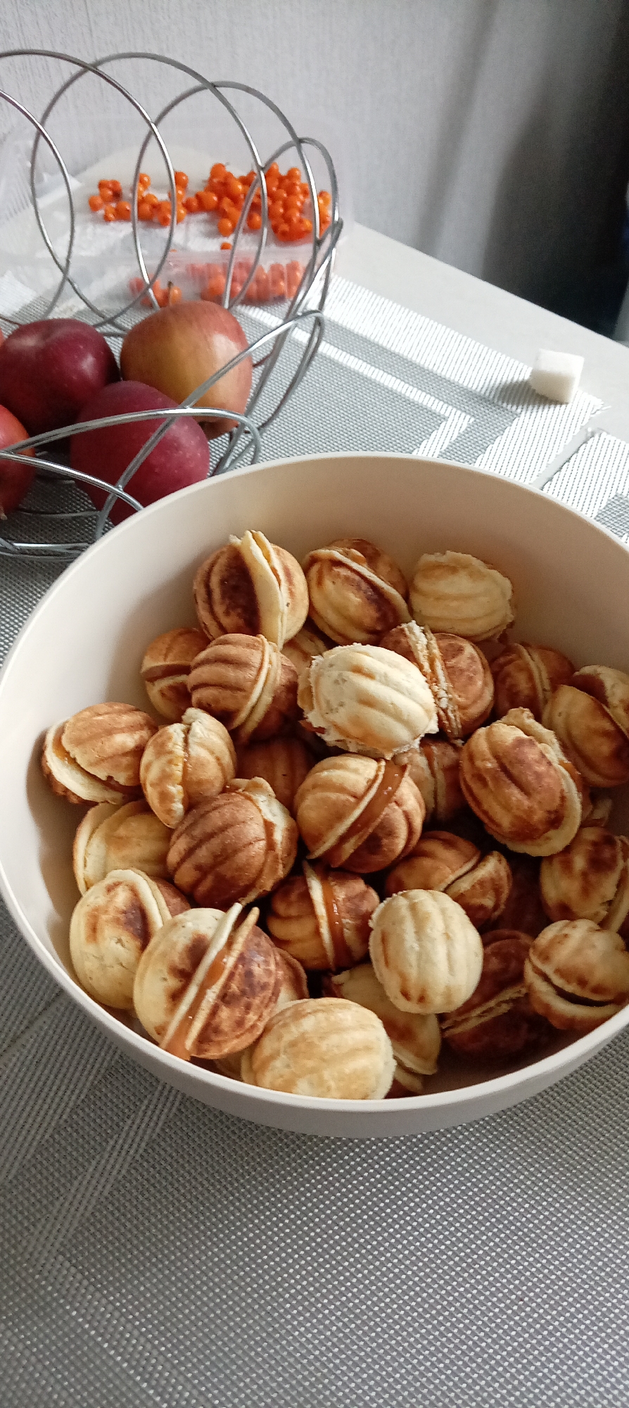 A high-angle, medium shot shows a bowl of walnut-shaped cookies, a wire basket with apples and rowan berries, and a sugar cube on a gray table with a grid pattern. The scene is likely indoors, possibly in a kitchen or dining area. The cookies are beige and brown, filled with what appears to be caramel. The apples are red, and the rowan berries are orange. The location is Chaikovsky, Russia. There are no people visible.