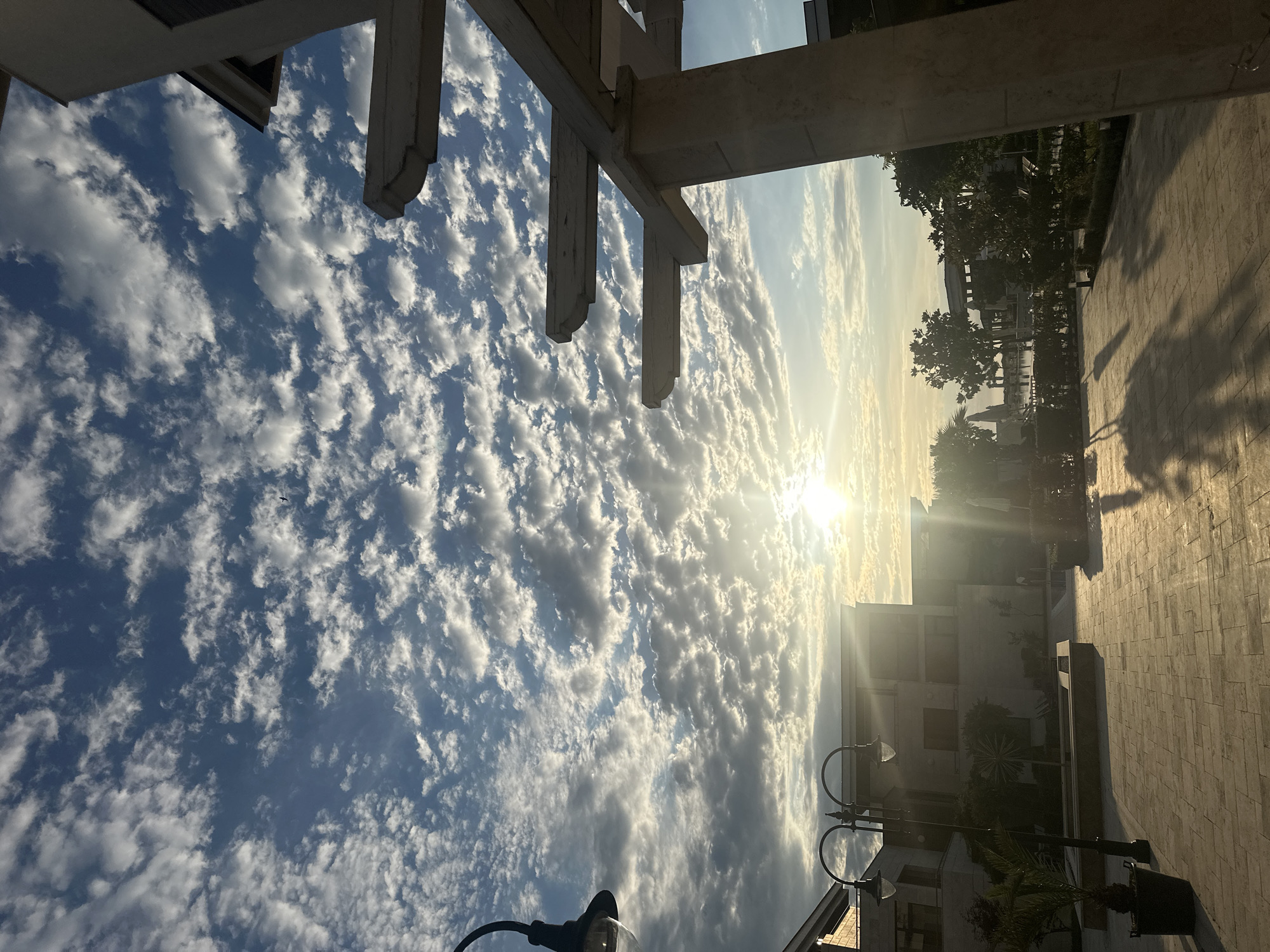 A low-angle outdoor shot captures a plaza area in Sozopol, Bulgaria, at what appears to be sunrise or sunset. The sky is filled with dramatic, textured clouds set against a bright blue backdrop. The sun is low on the horizon, casting a warm glow and creating long shadows across the paved plaza.
In the foreground, a beige stone pergola with wooden beams extends from the right side of the frame. To the left, a modern building with a beige facade is visible, featuring multiple windows. A black streetlamp with two lights stands near the building. Potted plants, including a palm tree, are placed around the plaza.
In the mid-ground, more modern buildings with similar beige facades are visible. Trees and lush greenery line what appears to be a waterfront or pool area. The water reflects the sky and buildings.
The overall impression is one of a serene and picturesque outdoor space, possibly a resort or a well-maintained public area, bathed in the soft light of a low sun.