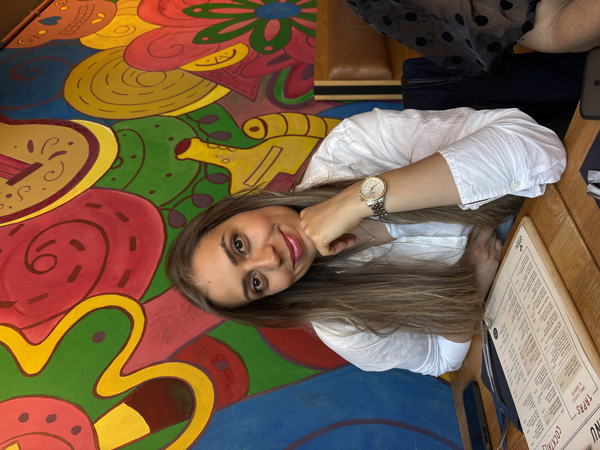 Here's a detailed description of the image:
The image shows a woman sitting at a table in what appears to be a restaurant or cafe. The scene is lit with natural light. The woman is fair-skinned with long brown hair, wearing a white button-down shirt and a wristwatch. She is looking at the camera with a slight smile and her hand is resting under her chin.
The table she is sitting at has a colorful, artistic design with painted elements. A menu is placed on the table, alongside cutlery, and a mobile phone. The menu advertises tapas and cocktails.
In the background, a person can be seen wearing a polka-dot shirt and sitting in a brown chair. The location seems to be in Luxembourg, Luxembourg as the menu on the table has a Luxembourg restaurant logo. The setting is a casual dining environment.