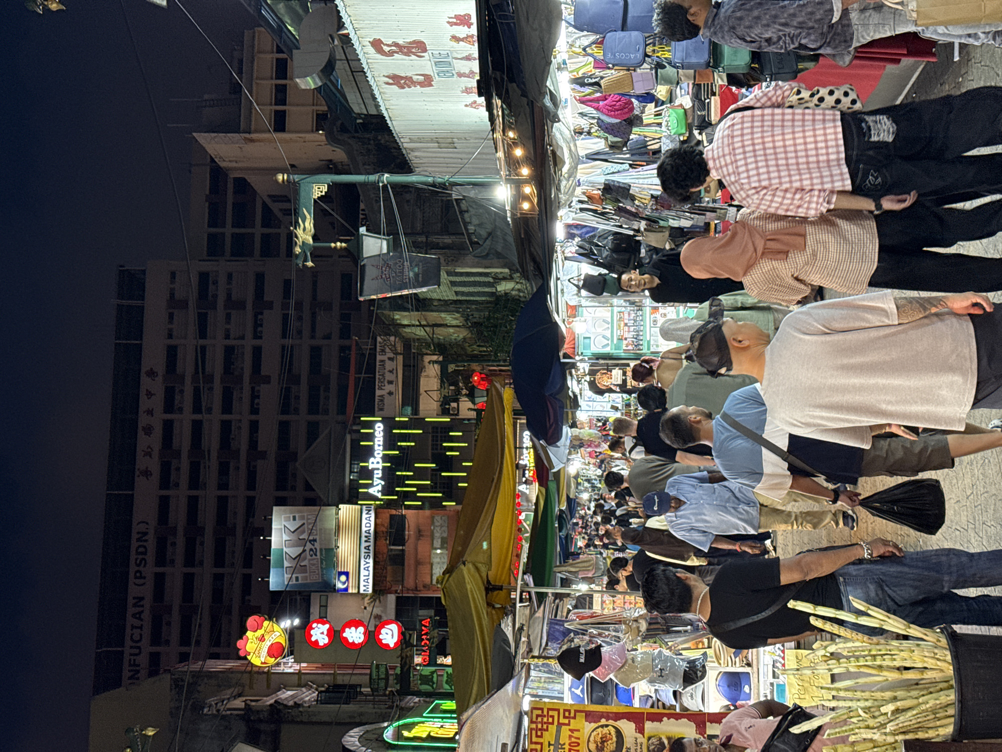 This photograph depicts a crowded street scene in Kuala Lumpur, Malaysia, captured at night. A narrow street, likely a pedestrian zone, is packed with people browsing merchandise displayed on stalls or blankets. The stalls appear to sell various goods, including clothing, bags, and accessories.
Overhead, a patchwork of awnings and signs shields the street. Chinese lanterns are hung, and neon signs illuminate the facades of buildings. The signage is predominantly in Chinese and English. Buildings are visible on either side, including a high-rise structure on the left with a sign reading "NFUCIAN (PSDN.)" alongside its Chinese translation. Another sign displays "MALAYSIA MADANI" and "Ayu Borneo."
Many people are visible, seemingly engaged in shopping or leisurely strolling. The street is well-lit, and despite the crowds, the atmosphere appears lively. A vendor is selling what looks like sugarcane sticks from a black plastic bucket.