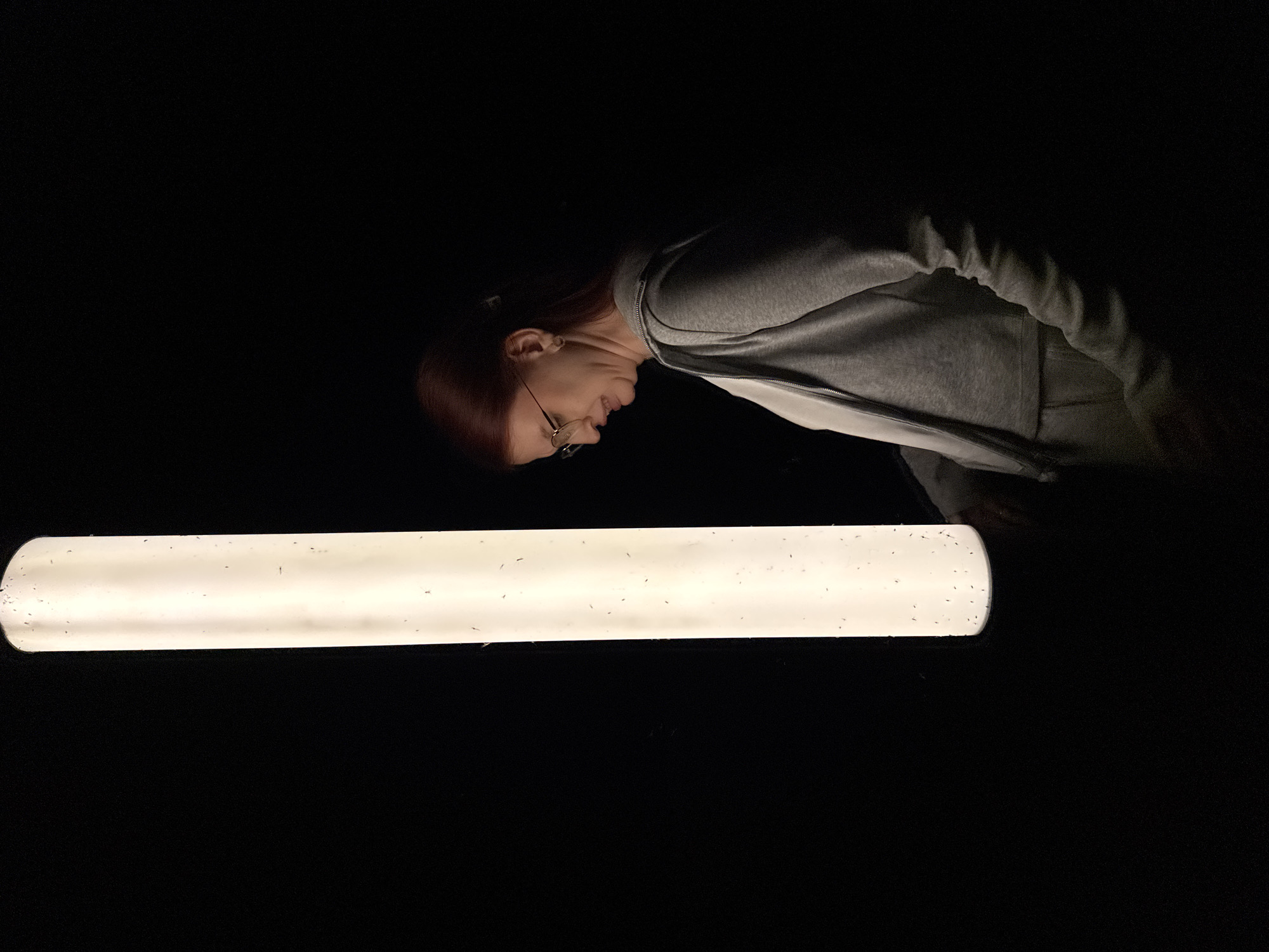 A woman with red hair and glasses wearing a gray hooded sweatshirt is bending over and looking at a long, white fluorescent light fixture in a dark room. The light fixture appears to be covered with small dark spots, likely insects. The location in the city of Nizhny Novgorod, Russia, is not possible to determine from the image.