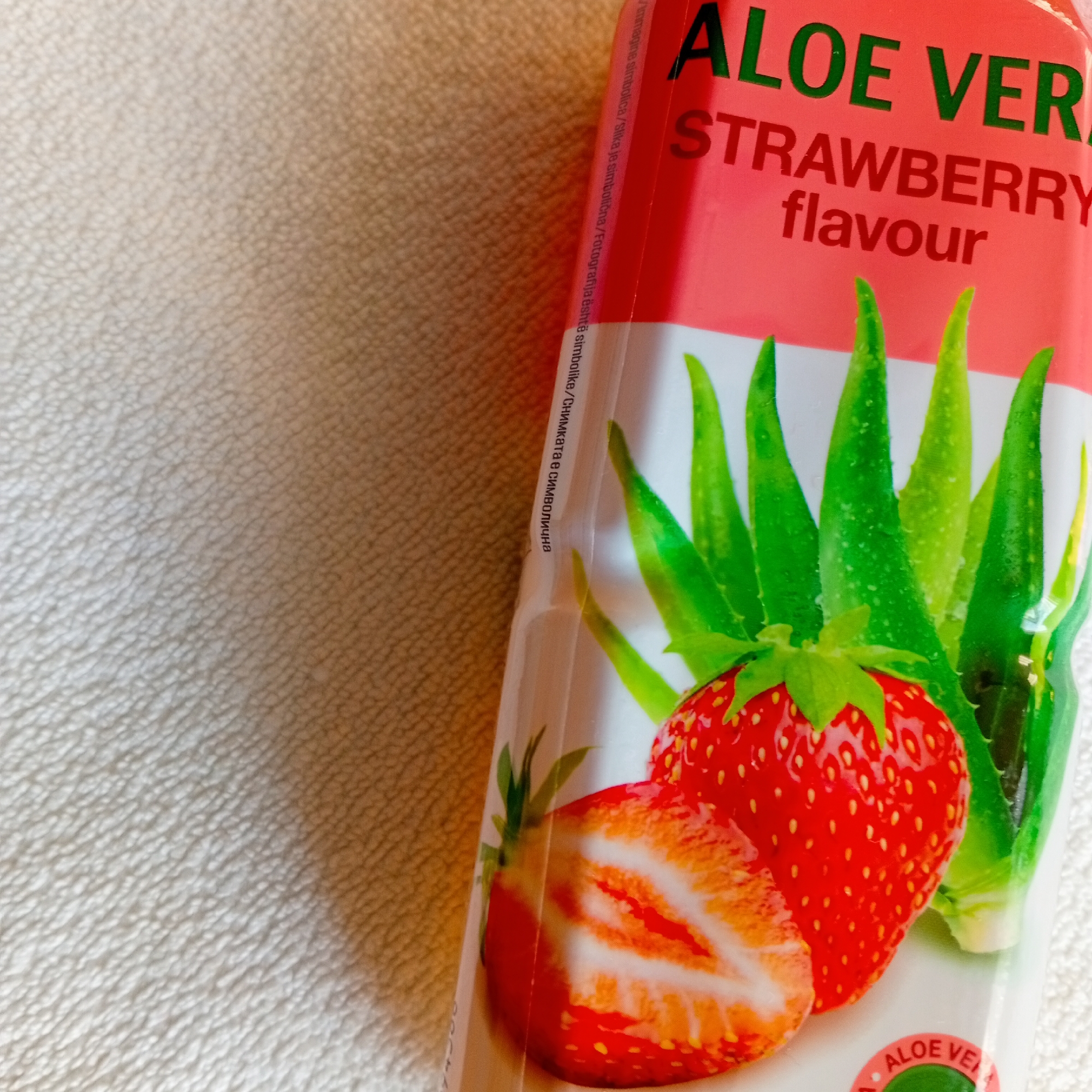 A close-up, angled overhead shot taken in Harmanli, Bulgaria, depicts a portion of a beverage container's label. The label prominently displays "ALOE VERA" in green text and "STRAWBERRY flavour" in red text against a red and white background. Below the text, an illustrative graphic features several green aloe vera leaves, a whole red strawberry, and a red strawberry sliced in half. Along the left edge of the label, small, vertically oriented text includes "Снимката е символична," indicating the image is symbolic. A partially visible circular logo with "ALOE VE" is located at the bottom right of the label. The container rests on a light beige, textured surface, likely fabric or upholstery, which fills the remainder of the frame, showing a subtle cross-hatch pattern and some tonal variation. No people, actions, or specific interactions are visible.