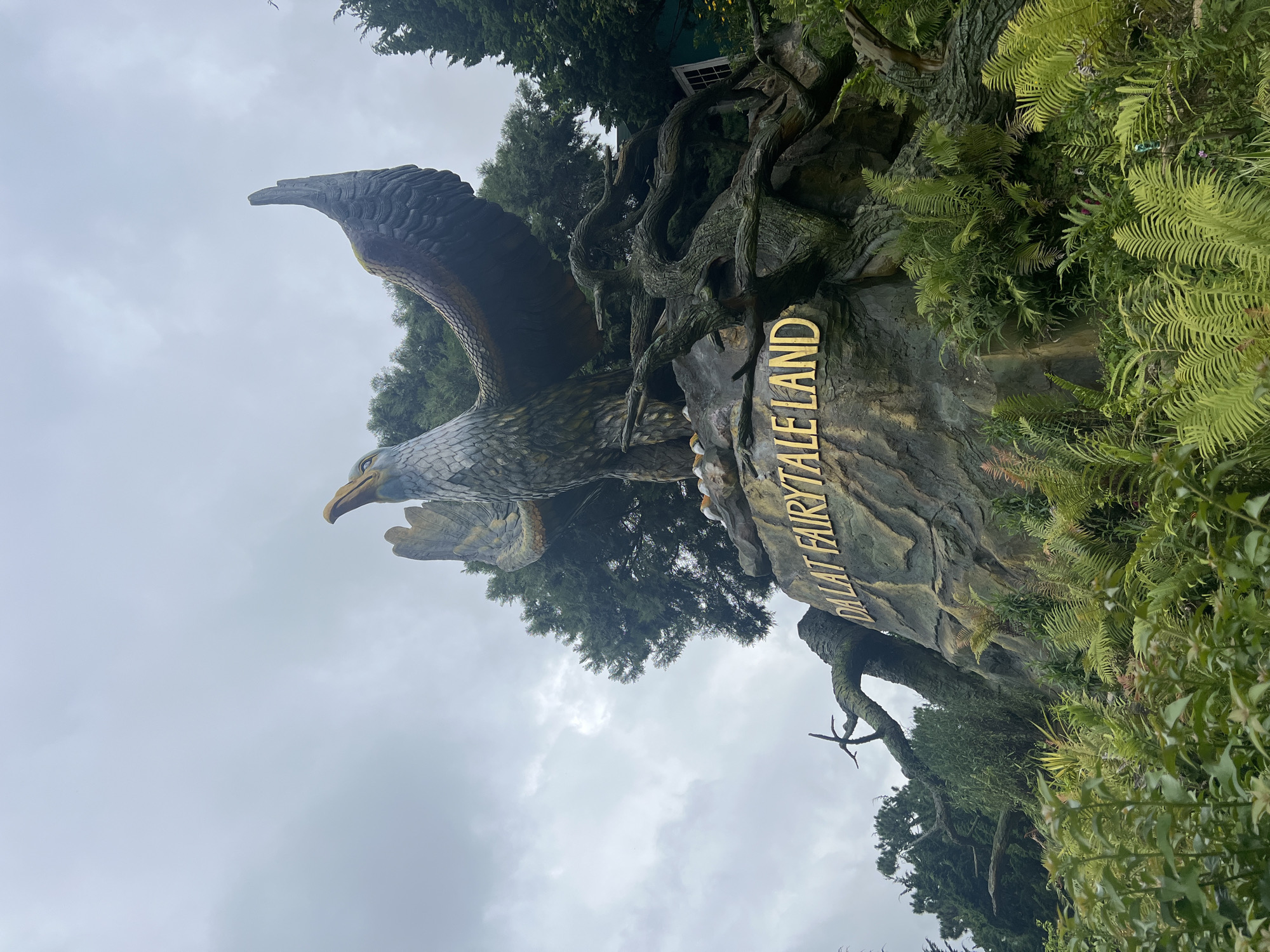 The image shows a large statue of an eagle perched on a rock formation, with its wings spread wide. The eagle statue is situated in an outdoor environment, surrounded by lush greenery and trees. The rock formation features the text "DALAT FAIRY TALE LAND" in large, golden lettering. The sky above is overcast with clouds. The statue appears to be an entrance or a prominent feature of the "Dalat Fairy Tale Land," which is a tourist attraction in Dalat, Vietnam, known for its whimsical and fairytale-themed decorations. The overall scene suggests a fantasy or magical theme.