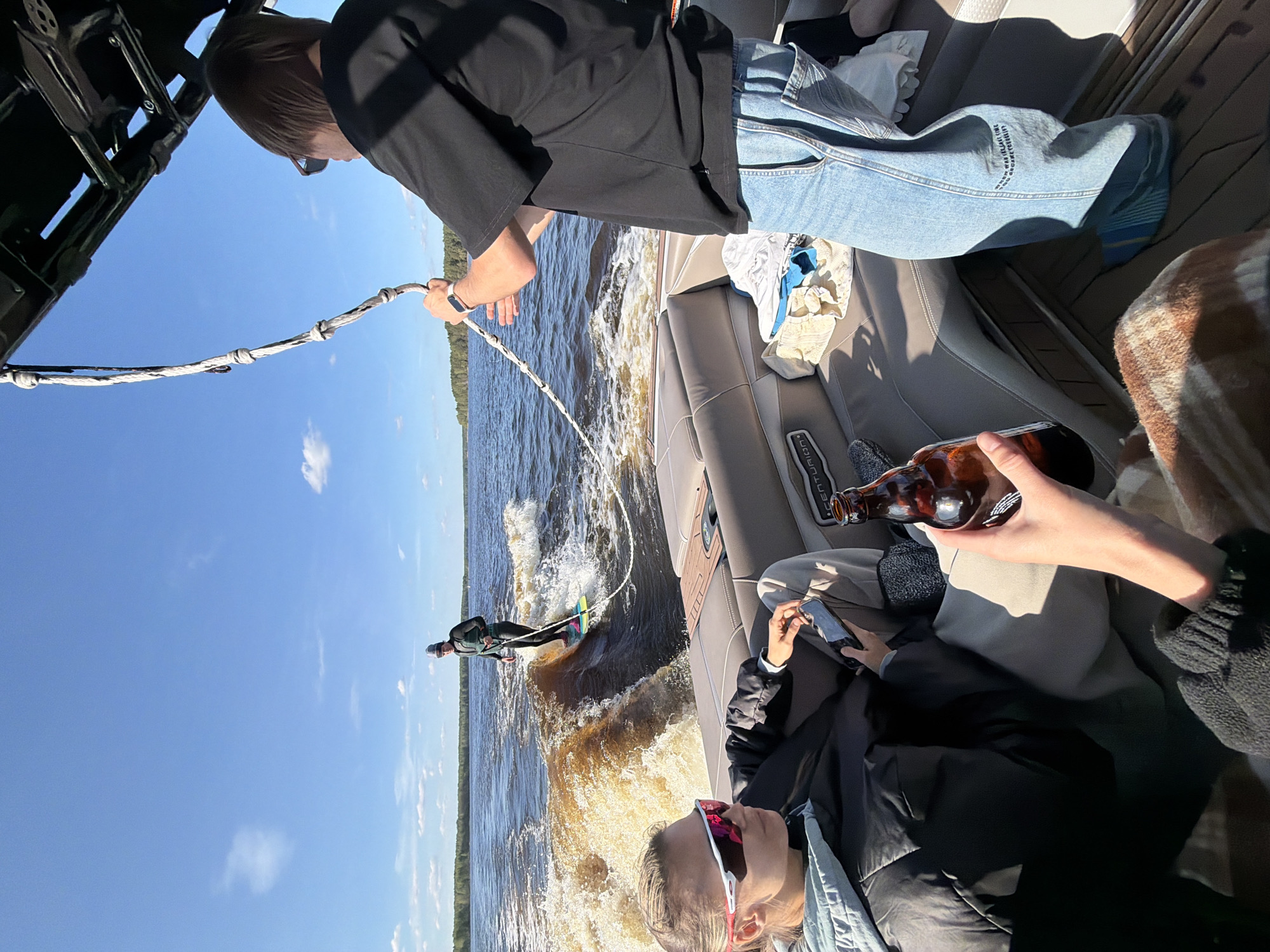 The image depicts a waterskiing scene, likely on a river or lake in Gorodishche, Russia, given the geotag. A person is waterskiing, being pulled by a boat. A person is operating the boat, holding the rope. In the boat, at least two other people are visible; one is holding a brown glass bottle and another person is operating a cellular device. The sky is clear and sunny. The water is choppy from the wake of the boat. The surrounding environment appears to be forested.