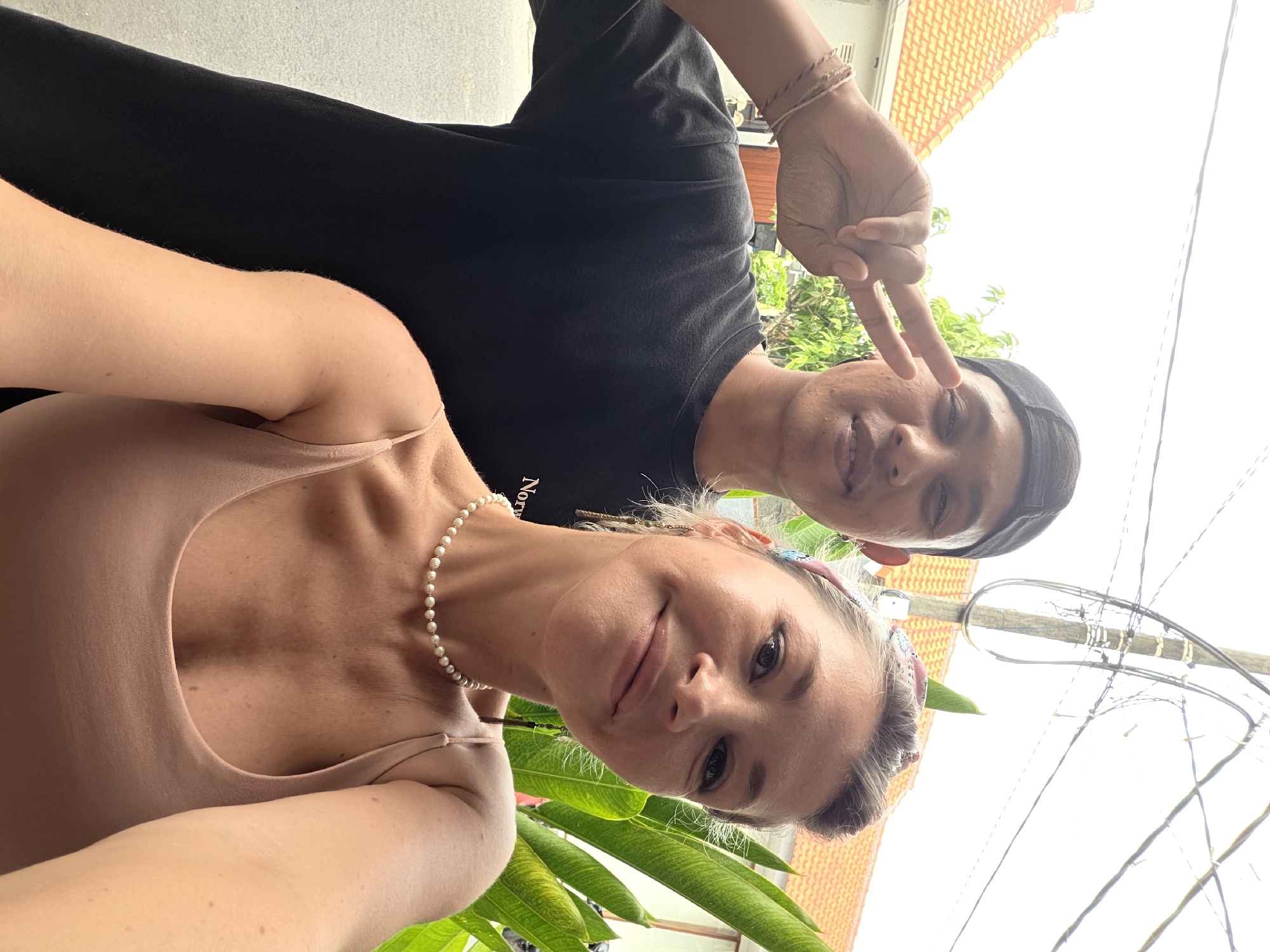 A medium shot photograph captures two individuals, a male and a female, posing for a selfie outdoors in North Kuta, Indonesia. The male, positioned slightly behind and to the left of the female, wears a black t-shirt and a black baseball cap. He is making a peace sign with his right hand, which is raised near his face. His left arm is bent at the elbow and resting on his side. The female, in the foreground, is looking towards the camera with a slight smile. She is wearing a light-colored tank top and a pearl necklace. Her right arm is extended, holding the camera for the selfie. Behind them, a tiled roof of a building is visible, along with some green foliage and utility wires overhead, suggesting an urban or suburban environment in Indonesia. The lighting suggests it is daytime, possibly overcast or late afternoon.