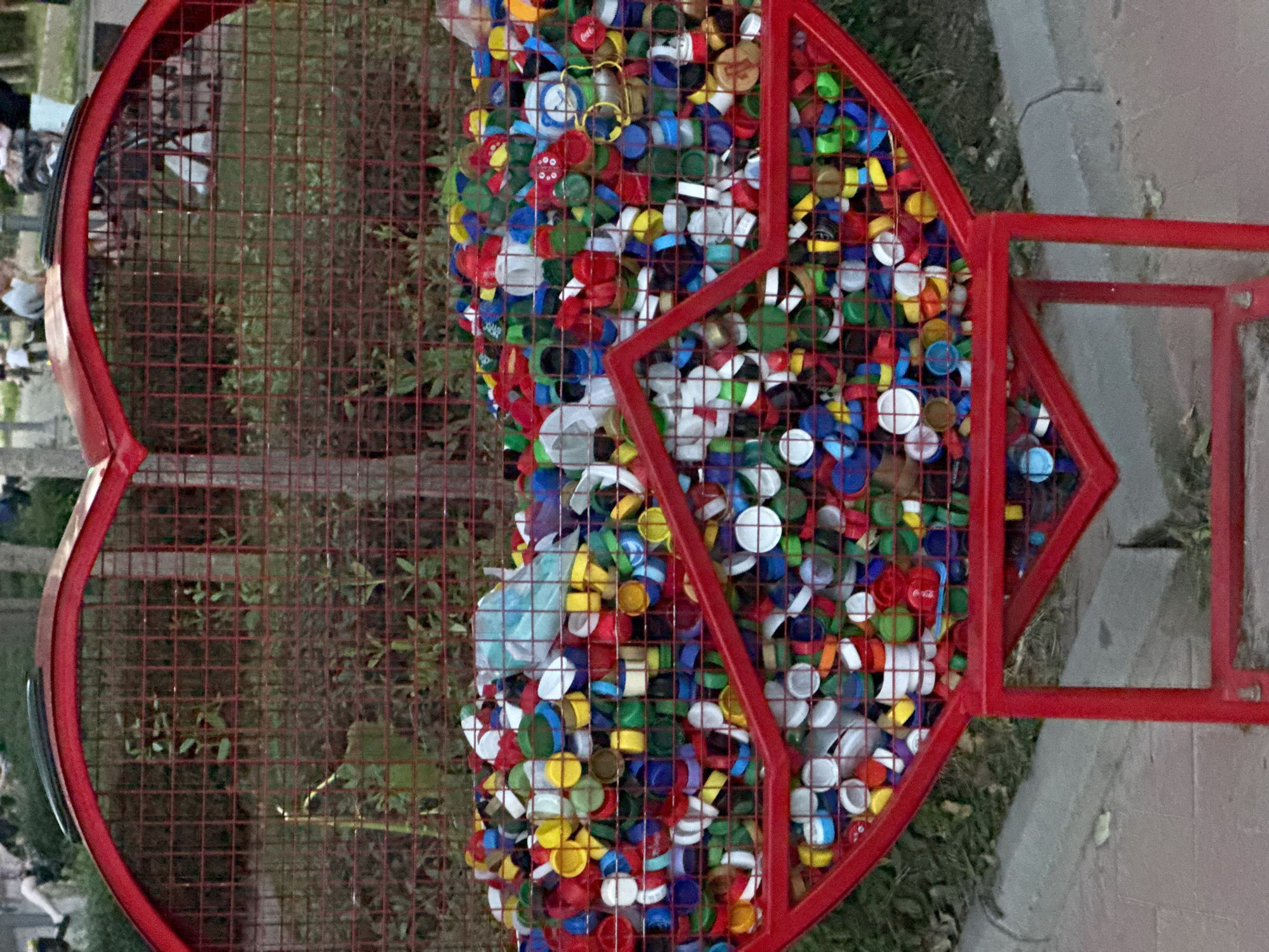 The image shows a large, red, heart-shaped metal trash receptacle filled with colorful plastic bottle caps. The receptacle is made of wire mesh and has a red frame. It is situated outdoors, with greenery and trees visible in the background. There are some people in the blurred background, but they are not the focus of the image. The bottle caps are of various colors, including red, blue, green, yellow, white, and black. Some caps appear to be from well-known brands, such as Coca-Cola. The receptacle itself is decorated with a red, zig-zag pattern formed by metal bars. The ground in front of the receptacle is paved with bricks and there is a concrete curb. The scene appears to be in a park or public space. The location is identified as the city of Kremenchuk, Ukraine.
