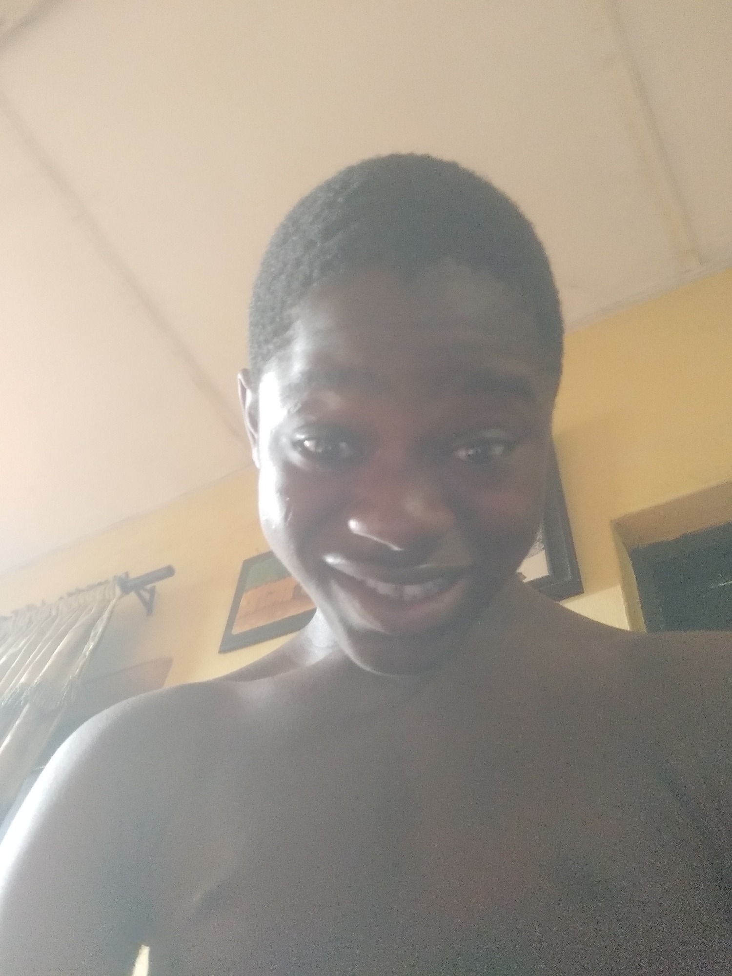 A low-angle, dimly lit close-up photo shows a young man with dark skin. He has short, dark hair and is smiling slightly, revealing his teeth. He appears to be shirtless. In the background, a tan-colored wall is visible, along with a partially obscured curtain, a framed picture, and a doorway. The location is likely indoors. There is no evidence to indicate it is in Shangisha, Nigeria.