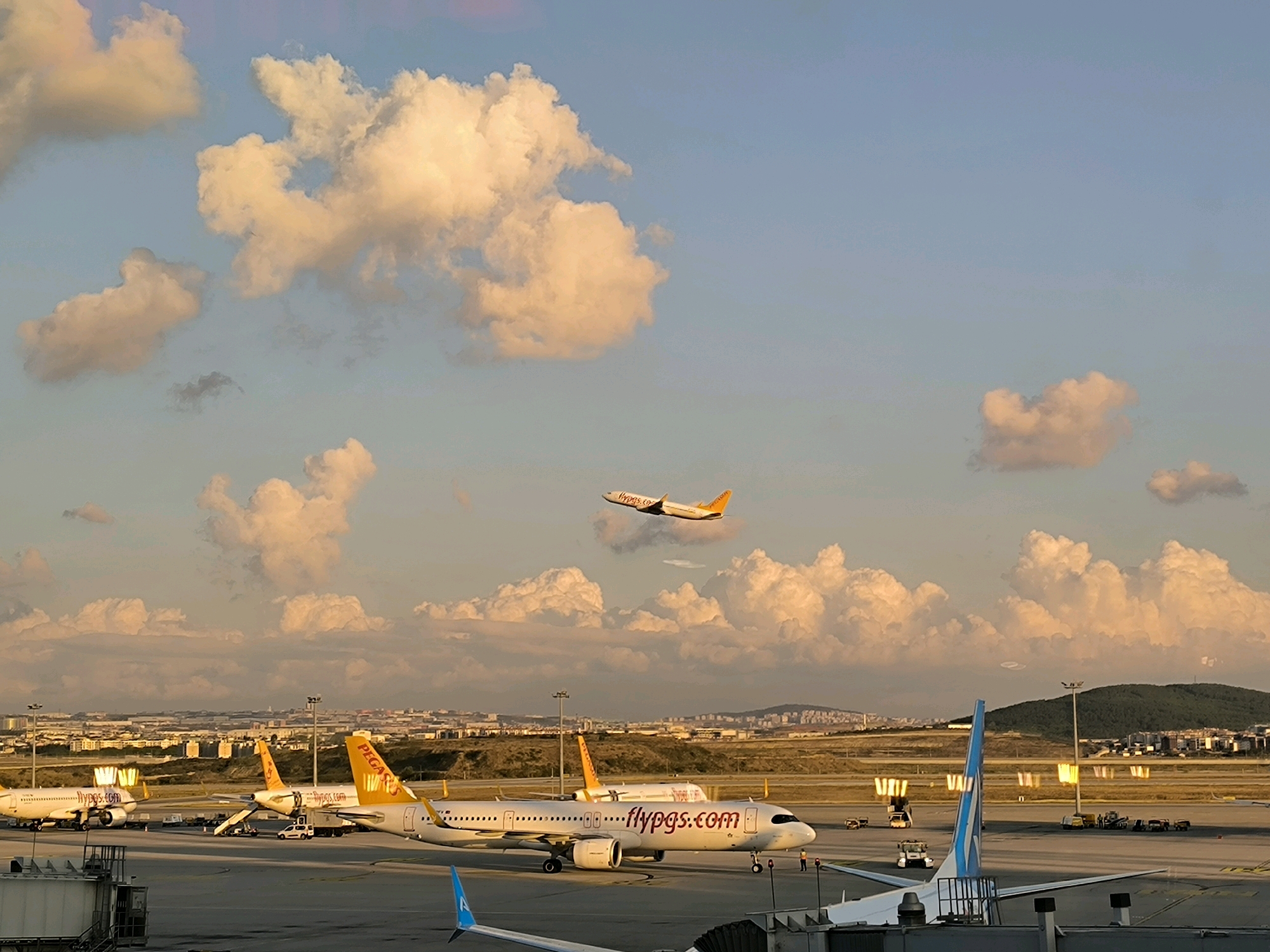 An airport tarmac at dusk in Pendik, Turkey, shows multiple Pegasus Airlines aircraft. In the foreground, a Pegasus Airlines airplane with the livery "flypgs.com" is parked on the tarmac. To the left of this plane, another Pegasus Airlines aircraft is visible. In the mid-ground, a third Pegasus Airlines plane is seen from the rear. High in the sky, a fourth Pegasus Airlines plane is in ascent, its landing gear retracted. The sky is a gradient of blue and orange, with large, fluffy clouds illuminated by the setting sun. In the distance, the cityscape of Pendik stretches out, bordered by hills. The scene is an active airport environment with service vehicles and airport personnel visible on the tarmac.
