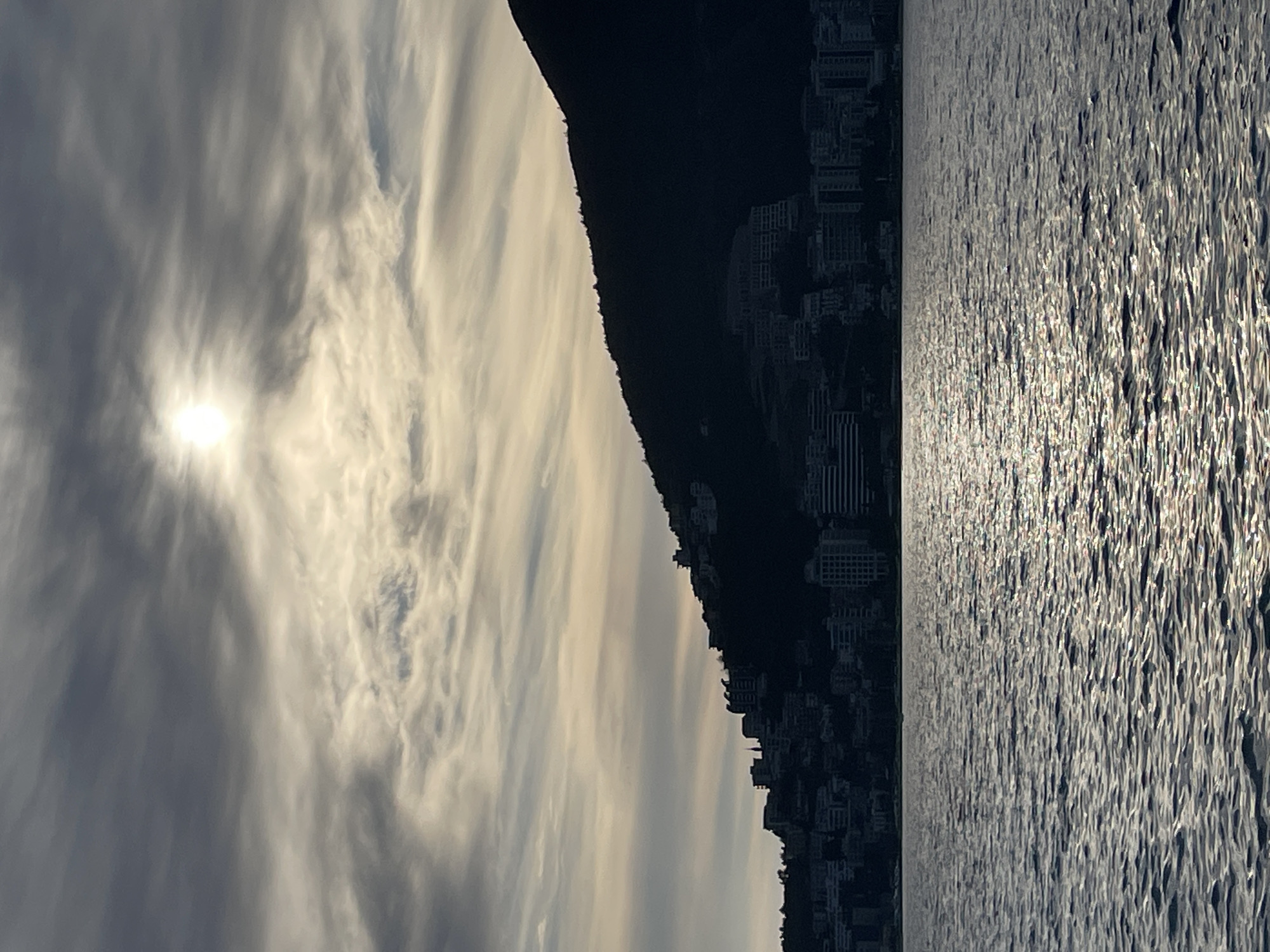 The scene depicts a coastal view in Budva, Montenegro. The foreground consists of a body of water with small, numerous waves on its surface, reflecting sunlight in scattered highlights across the ripples. In the midground, a large, dark mountain or hill rises directly from the water, silhouetted against the sky. Along the lower slopes and at the base of this mountain, multiple multi-story buildings are visible, also appearing as dark silhouettes against the lighter sky. A slender spire structure is discernible among the buildings on the left side of the mountain's base. The sky occupies the upper portion of the image, featuring a mix of elongated, streaky clouds in shades of grey and pale yellow. The sun, appearing as a bright, diffuse disc, is positioned in the upper-left quadrant of the sky, providing backlight to the scene. No individuals or specific actions are observable.