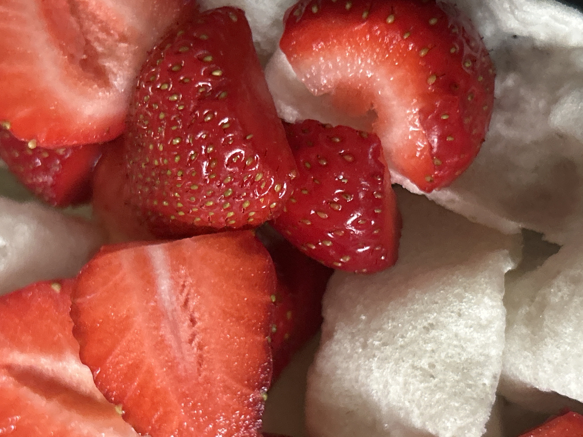 The image shows a close-up of sliced strawberries mixed with white, porous material. The strawberries are bright red and show their yellow seeds. The white material has a rough, uneven texture, suggesting it might be a type of foam or packing material. The location is Unknown, Unknown.
