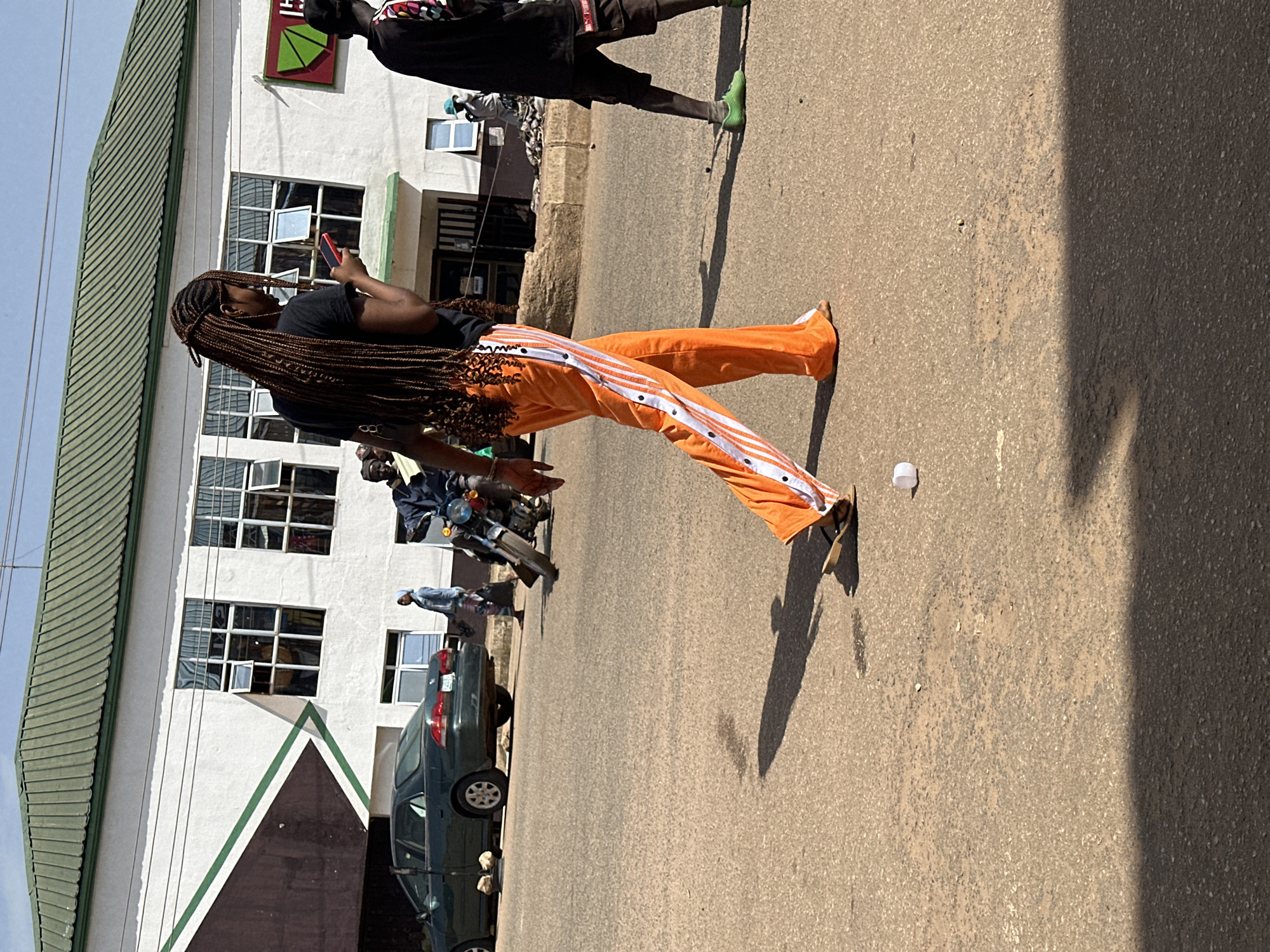 In Bukuru, Nigeria, on a sunny day, a young woman with long, braided hair walks across a paved road, engrossed in her red smartphone. She is wearing a black t-shirt and vibrant orange sweatpants adorned with white stripes and silver snaps down the sides. Her footwear appears to be flip-flops or sandals. To her right, another person, partially visible, walks away from the camera wearing a black t-shirt, dark shorts, and green shoes. In the background, a dark green sedan is parked on the side of the road. Further behind, a building with a green-tiled roof and white walls features several windows. A motorcycle with a rider is also visible in the middle ground, along with other pedestrians. The road surface is asphalt, showing signs of wear. A discarded white cup lies on the road near the main subject.