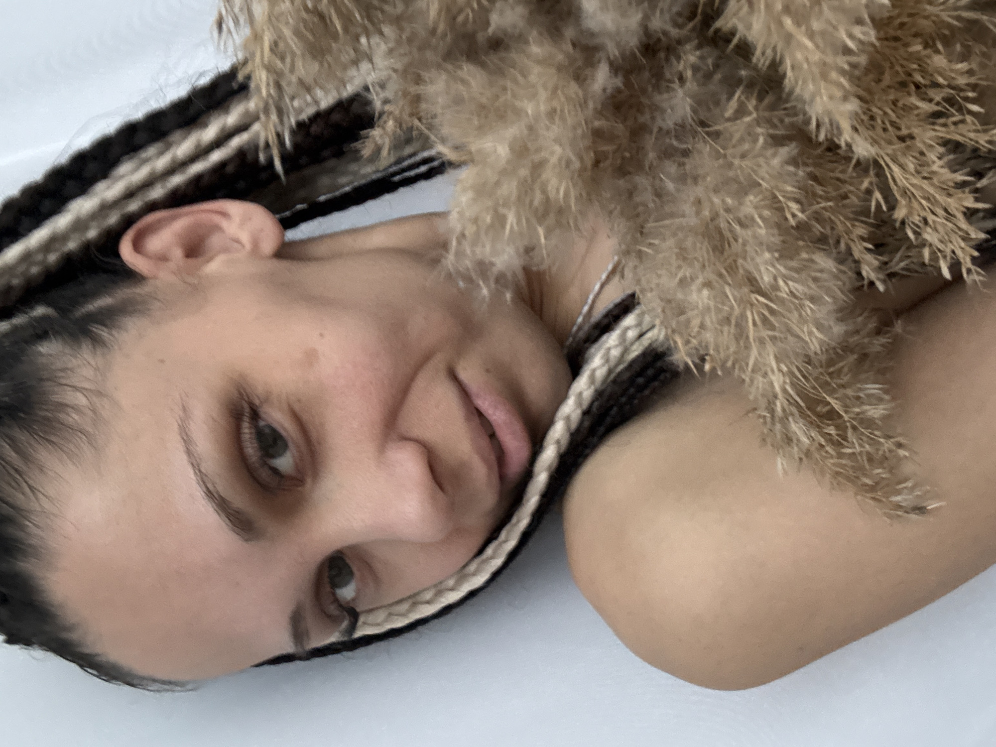 This close-up, rotated portrait captures a person lying on a light-colored surface in Rusanovo, Russia. The subject is a person with natural skin tone and dark hair styled in numerous thin braids, accented with lighter, possibly blonde or cream-colored, sections. She looks directly at the viewer with a subtle, gentle smile or smirk on her lips, her gaze steady and light-colored eyes slightly narrowed.
A large bundle of dried, feathery, light brown plants, resembling pampas grass or similar ornamental grasses, is draped over her right shoulder and neck, extending towards the upper right of the frame. A delicate silver chain necklace is visible around her neck. Her left ear is partially seen, and her facial features appear natural, suggesting minimal or no makeup. The person's bare shoulders and arm are visible, resting against the plain, light-colored background. The lighting is soft and diffused, creating a serene and intimate atmosphere. No visible text is present in the image.