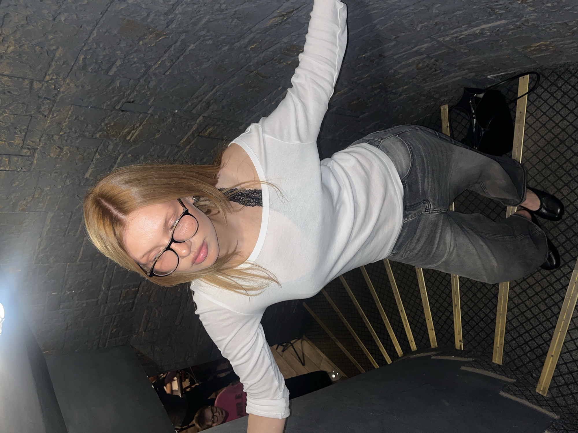 The image shows a young woman with blonde hair and glasses, wearing a white long-sleeved shirt and gray jeans. She is posing for a photo while standing on a carpeted staircase. A black handbag is visible on the side of the stairs. The staircase has wooden railings and a patterned carpet. The background appears to be an indoor setting with textured stone walls. The lighting suggests it might be evening or indoors. The woman's expression is neutral, and the overall mood is casual. There is no visible text in the image. The context indicates the location is Chaikovsky, Russia.