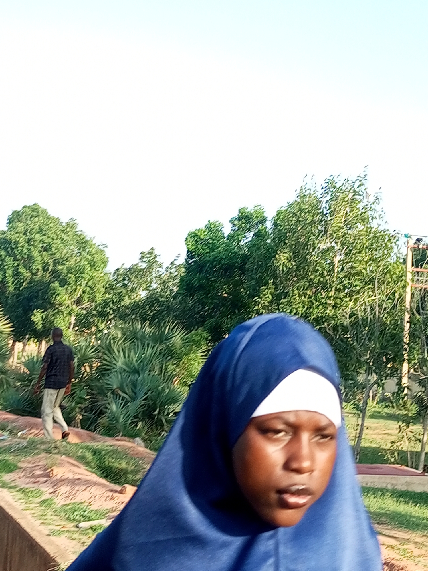 The image depicts a scene in Garko, Nigeria, featuring a young woman in the foreground wearing a blue hijab and a white underscarf. She has dark skin and is looking to her right, with her mouth slightly open. Behind her, a man in a black shirt and light-colored pants walks away from the camera, his back partially visible. The environment consists of lush green trees and foliage, with a bright, clear sky in the background. There are also some man-made structures, including what appears to be a fence or scaffolding on the right side of the frame. The ground is covered with a mix of dirt, sand, and grass, suggesting an outdoor setting, possibly a park or a public area.