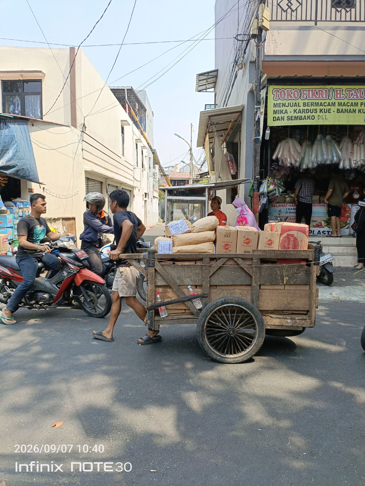 In a street scene in Central Jakarta, Indonesia, a man pushes a wooden cart laden with boxes and sacks. The cart has two large spoked wheels at the rear and is positioned on an asphalt road. To the left, two men on motorcycles are engaged in conversation. The motorcycle on the left is red, and the one behind it is black. A building with cream-colored walls and dark windows is visible in the background on the left, while on the right, a shop with a yellow banner reading "TOKO FIKRI / H. TAU" is open, displaying various goods. The banner also lists items like "MENJUAL BERMACAM MACAM PERLENGKAPAN MIKA - KARDUS KUE - BAHAN KUE DAN SEMBAKO DLL". Several people are seen inside and in front of the shop. The sky is clear and bright, suggesting daytime. The scene captures a moment of daily commerce and transportation on a city street.